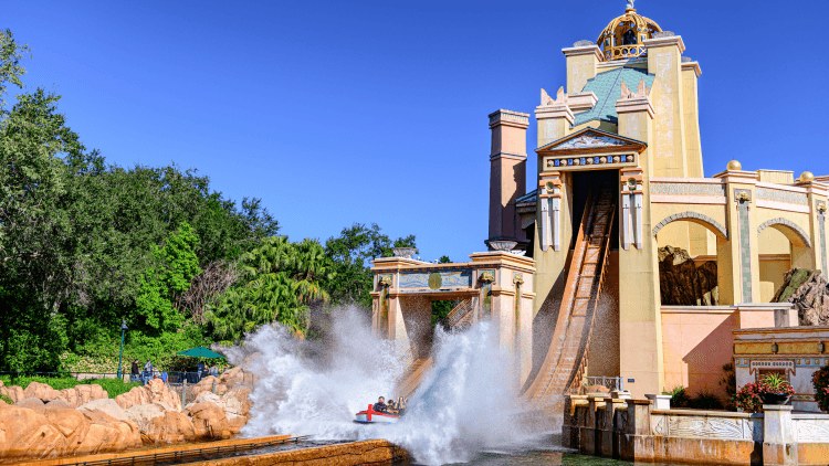 A wide daylight view of the Journey to Atlantis facade under a cloudless blue sky. A pale stone tower with a blue and gold cupola stands at right beside the steep brown flume drop, and a red boat of riders hits the splashdown in a tall wall of white spray, with green trees and rockwork at left