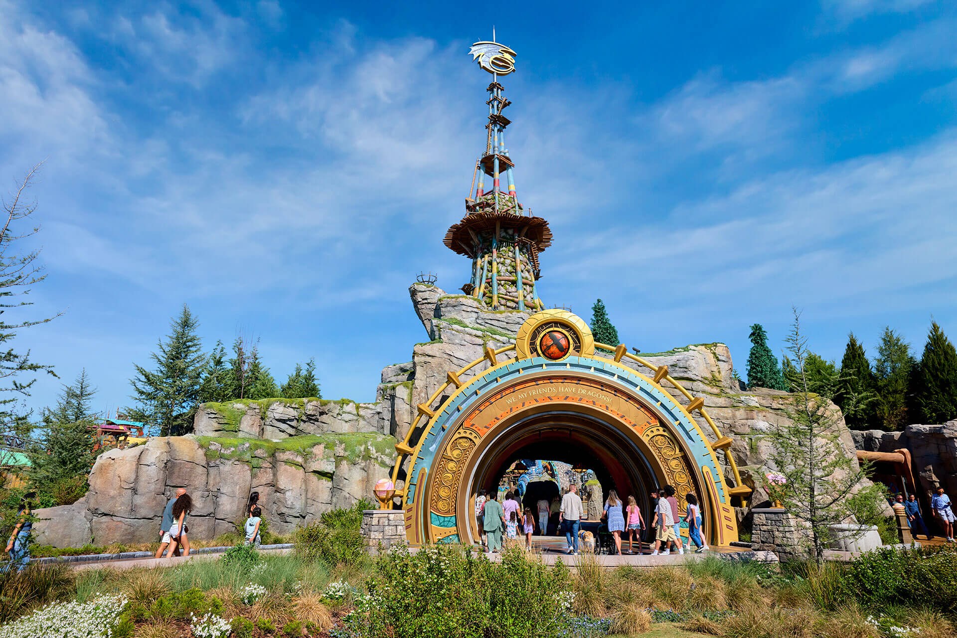 The Isle of Berk entrance portal, a large gold and teal circular archway set into rockwork lettered WE, MY FRIENDS, HAVE DRAGONS! along its inner band, with guests walking through it, a tall stone and timber watchtower topped by a metal dragon emblem rising behind, and open blue sky above
