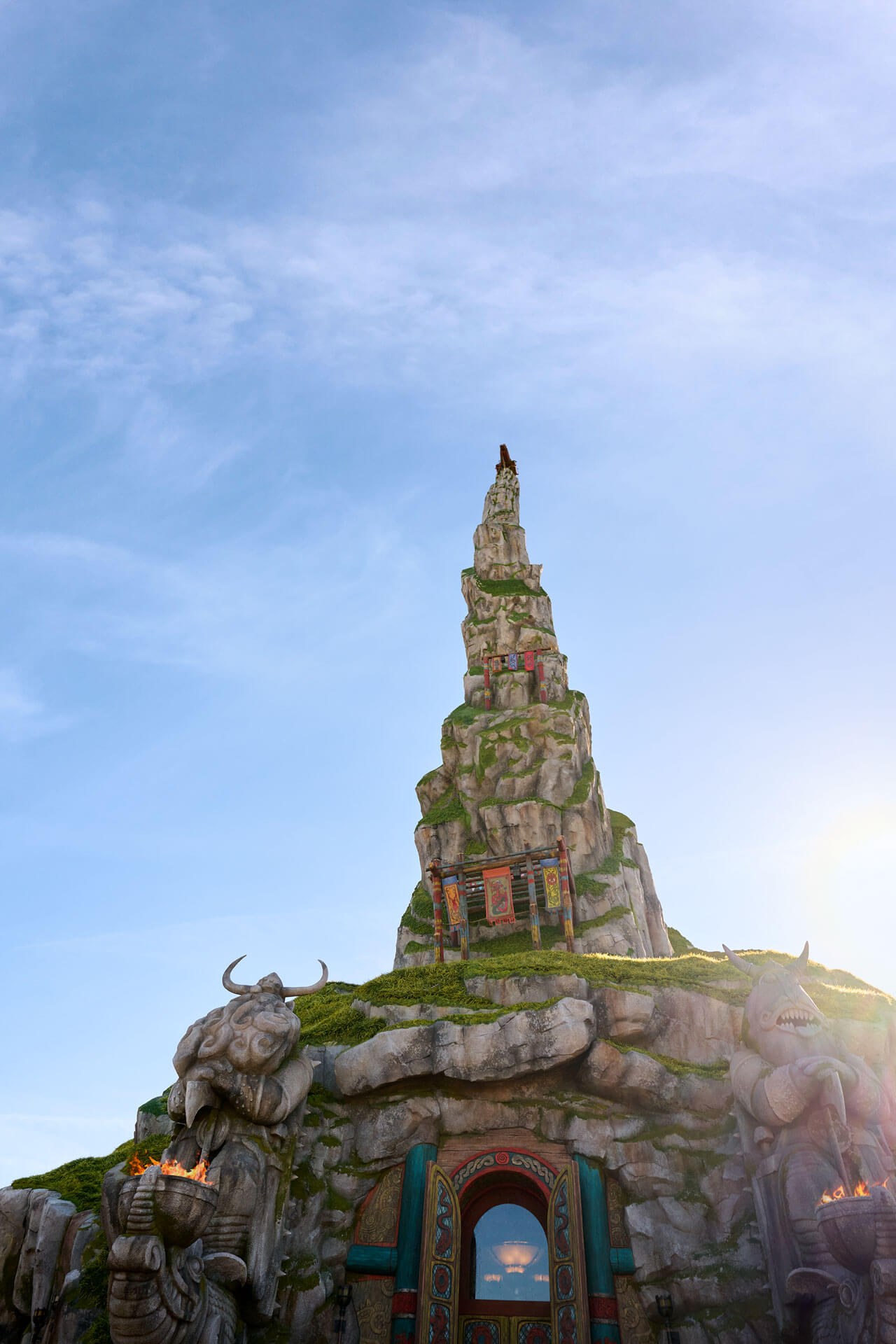 A low upward angle view of the tall craggy rock spire at Isle of Berk, moss covered and hung with painted banners, filling the lower two thirds of a tall frame under a wide pale blue sky. Carved stone Viking statues with lit fire braziers flank an ornate teal and gold doorway at the bottom