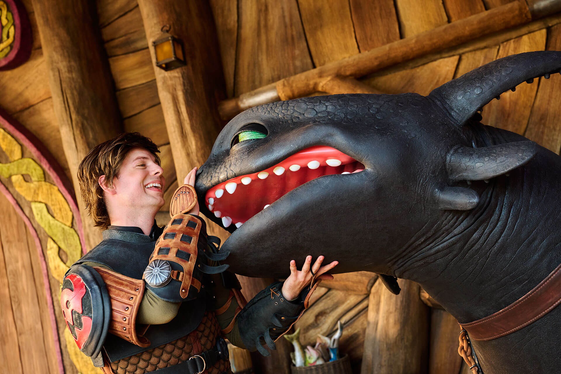 Inside a timber Viking hall, the Hiccup character performer in leather armor laughs with one gloved hand on the snout of a large black Toothless figure and the other hand under its jaw, its mouth open showing white teeth and a red interior, its one visible eye green