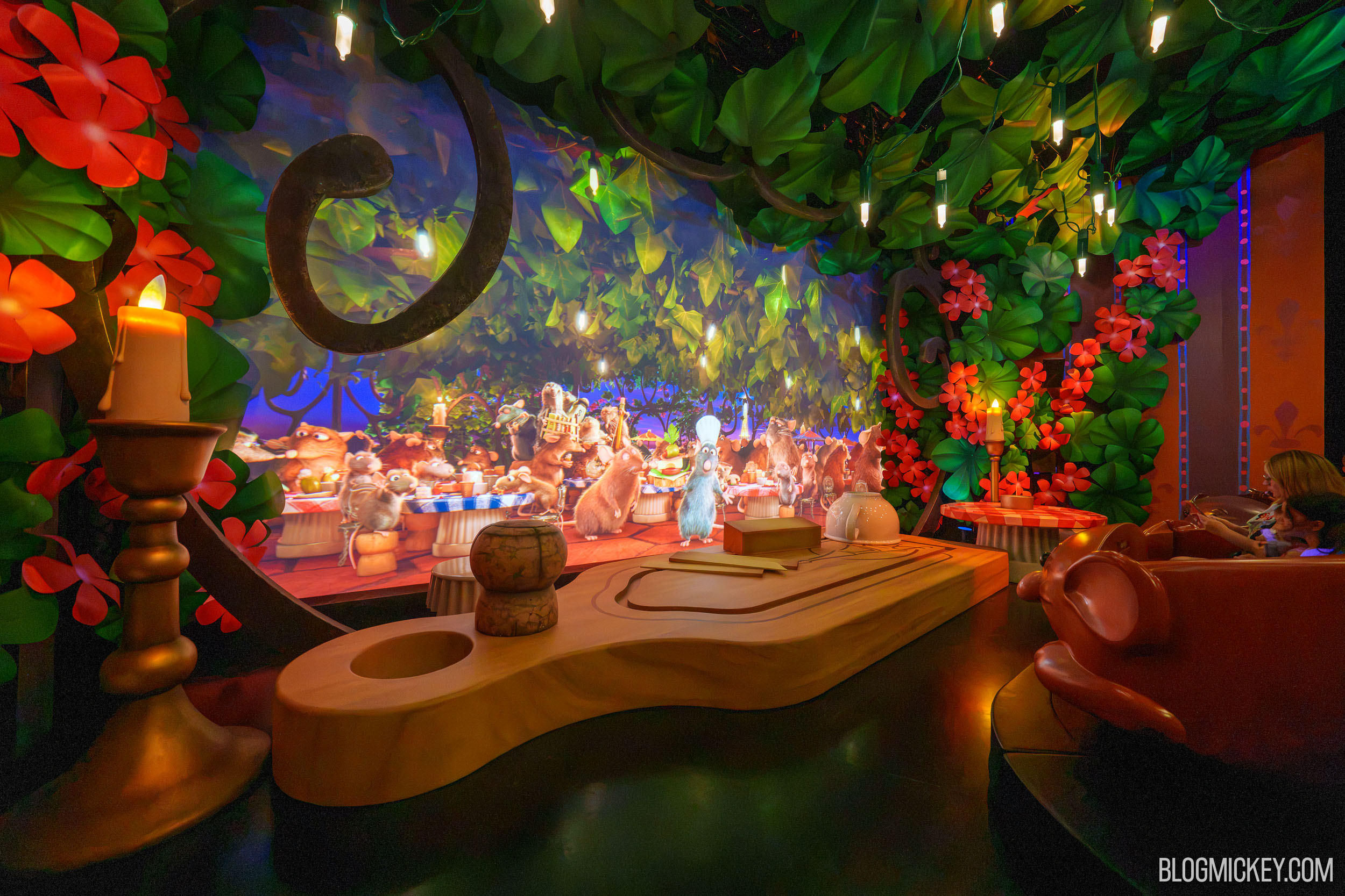 An on-ride view of the rooftop finale scene. A large pale wood cutting board fills the foreground, holding a rounded cork stopper, a flat tan block and an upturned white ceramic pot. Behind it a screen shows dozens of rats feasting at checkered tables with Remy in his chef's toque at the center. Oversized red flowers, green leaves and a lit candle on a gold candlestick stand around the screen as physical set pieces