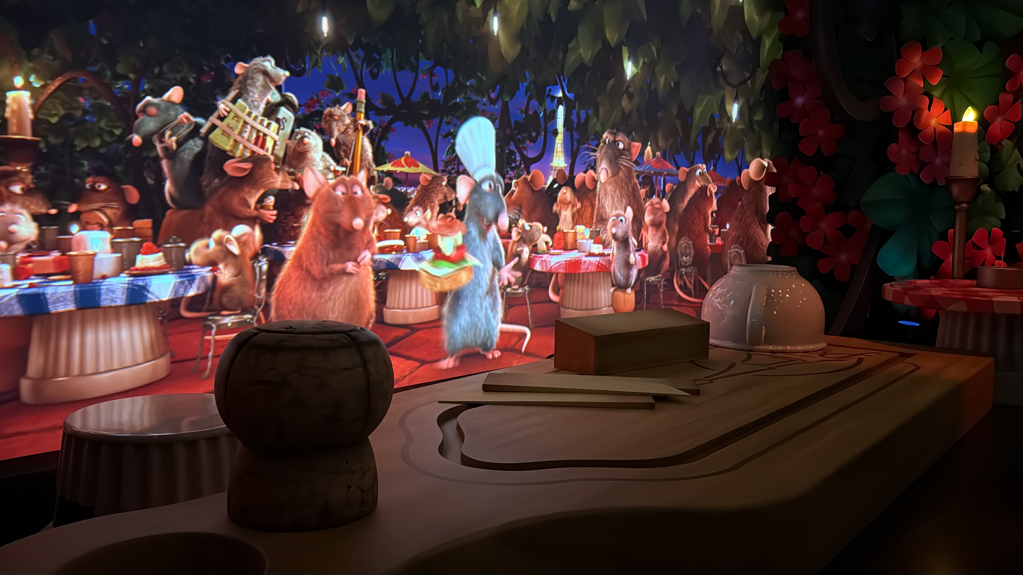 A closer view of the same finale scene. The cutting board fills the lower right of the frame, with a squat mottled cork sitting at its left end, a tan block and an upturned white pot beside it. The screen behind shows the rat banquet, Remy standing at center in a chef's toque, and the Eiffel Tower against a blue dusk sky