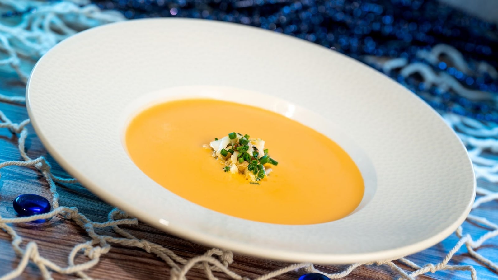 A wide rimmed white bowl holding a smooth orange bisque topped with a small pile of white seafood and chopped chives, resting on a wooden surface with fishing net and blue glass beads around it