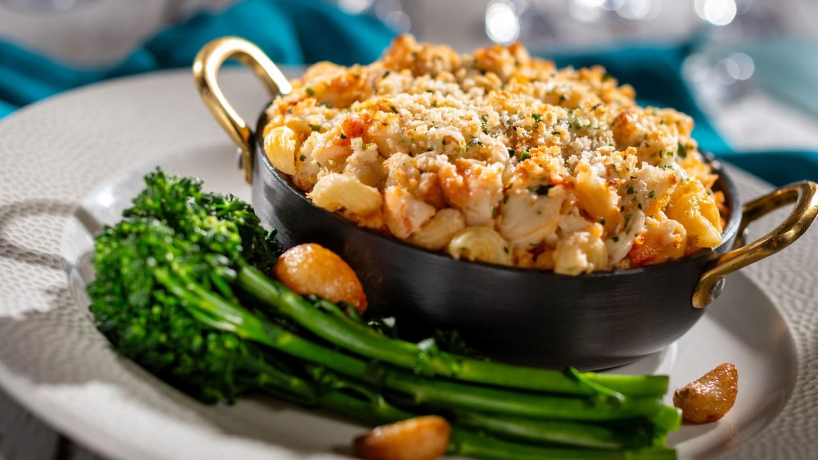 A black cast iron pan with brass handles holding the Seafood Mac and Cheese under a browned breadcrumb crust, plated with broccoli and roasted garlic cloves on a white dish
