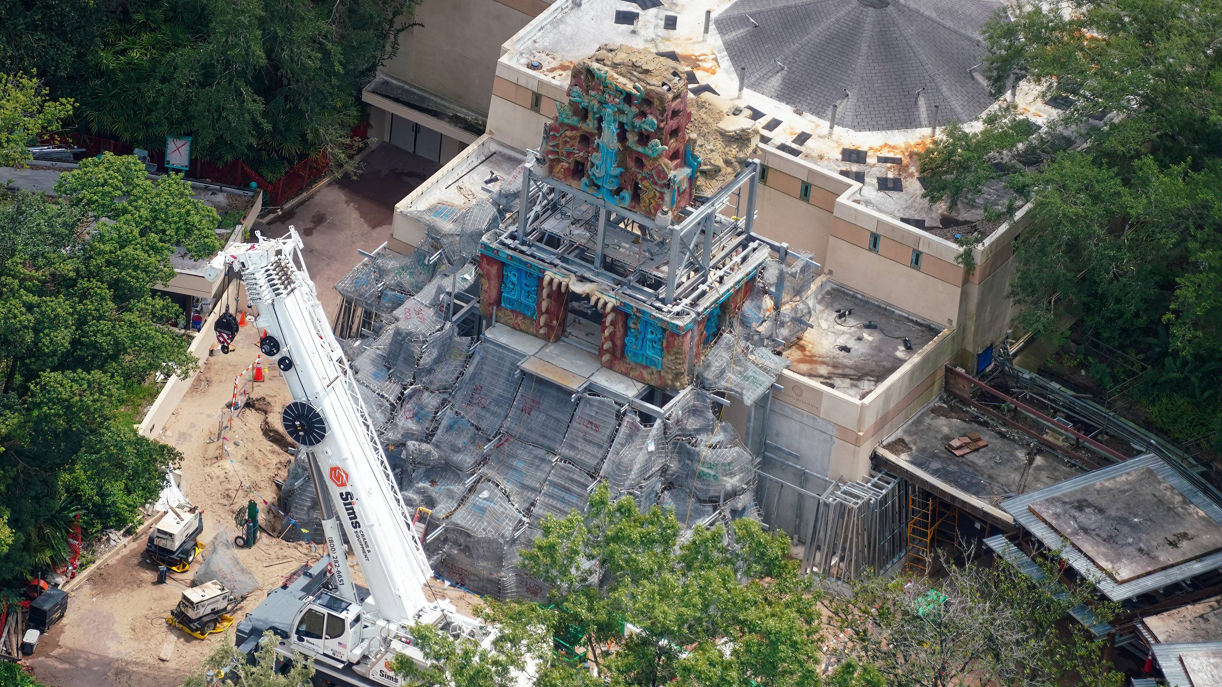 A wider aerial of the same construction site. A white Sims Crane and Equipment mobile crane stands at the left with its boom extended away from the temple, its hook block hanging over a sandy backstage service area beside a low beige building. The partly built pyramid rises at center with painted red and turquoise carved sections at its top and around its lower level, separated by exposed gray steel framing. Broad sloping pyramid steps below are covered in gray wire mesh marked with red and blue spray paint. Trees, sand and construction fencing surround the site