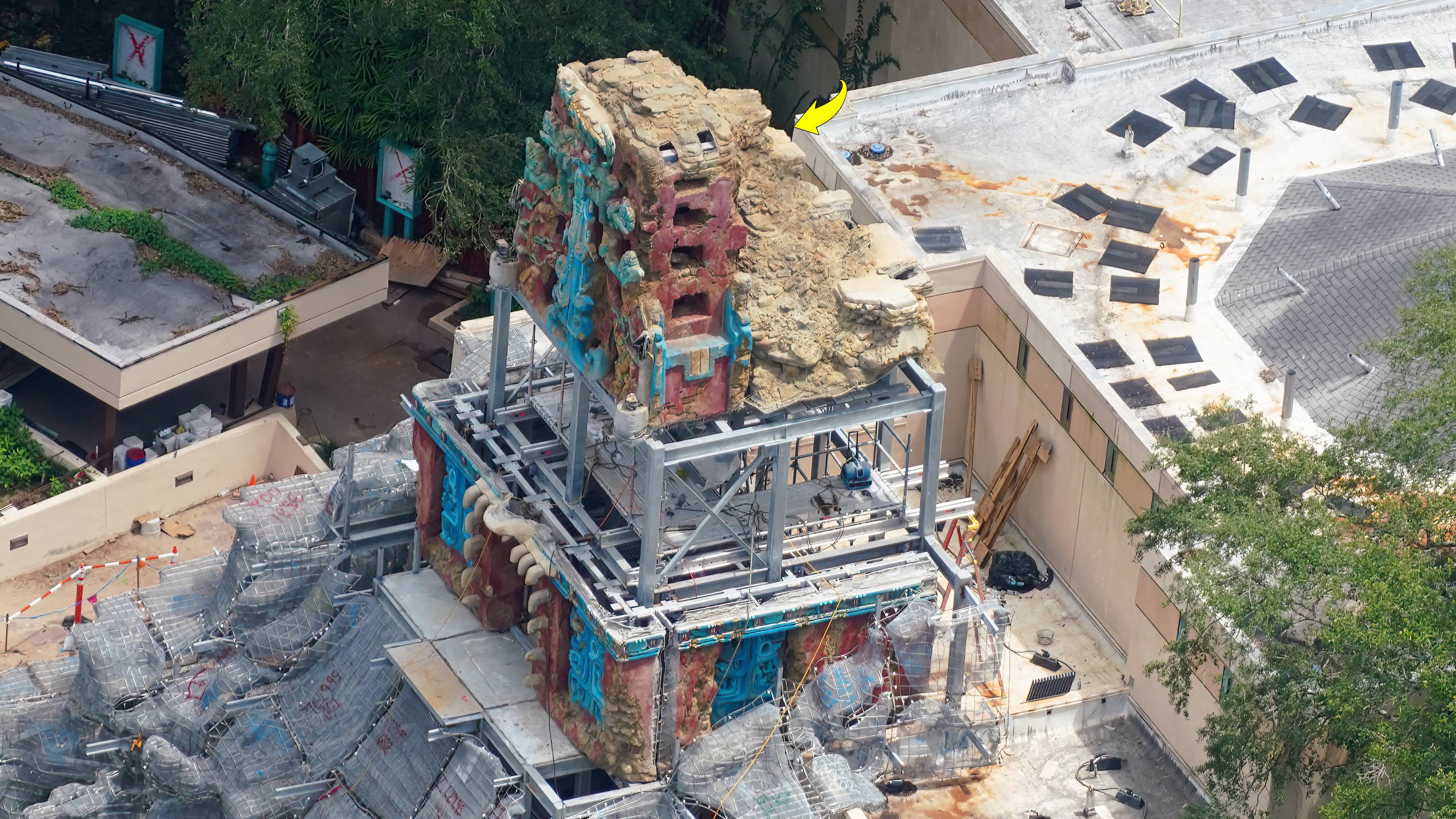 An oblique aerial view of the Indiana Jones show building. A tall finished temple section sits at the top, carved with turquoise scrollwork and deep red panels pierced by dark rectangular openings, its upper edge trailing off into pale tan crumbling rock. Below it stands an open gray structural steel frame with nothing on it. Beneath that sits a second finished section in the same red and turquoise, with a row of pale stone teeth along its edge. Gray wire mesh rockwork spreads out around the base and a yellow arrow points at the top of the upper section