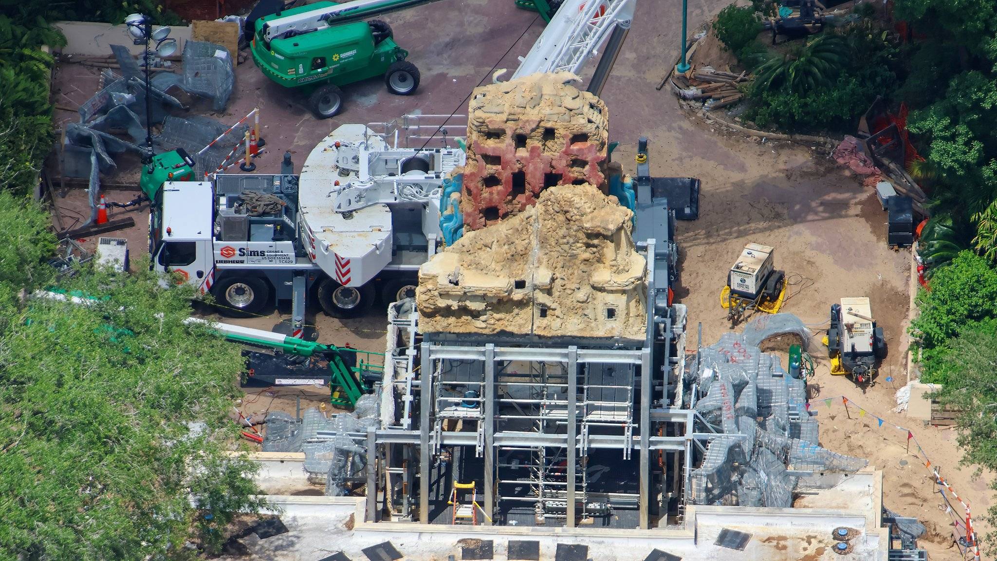 An oblique aerial of the temple dominated by sculpted pale tan rock carved into stratified crumbling ledges. A faded red panel pierced by a grid of dark rectangular openings sits in the middle of the structure, with bright turquoise carved bands flanking it on both sides. A white Sims Crane and Equipment truck crane is parked at the left beside a green boom lift, and open gray steel framing and scaffolding stand exposed at the base of the structure