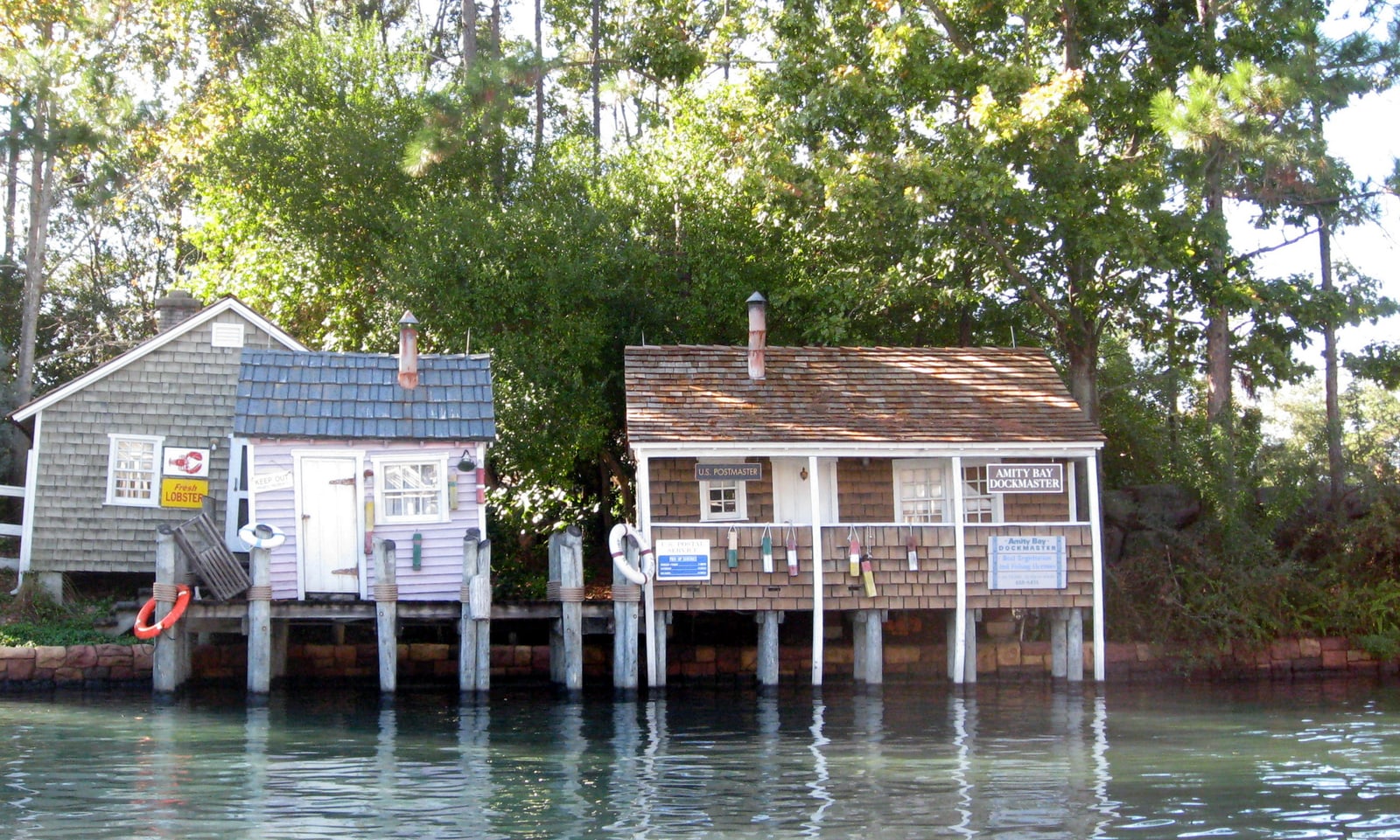 Two small weathered shingle buildings standing on wooden pilings over green water, one with signs reading U.S. Postmaster and Amity Bay Dockmaster, the other pale pink with a Fresh Lobster sign on the grey building behind it