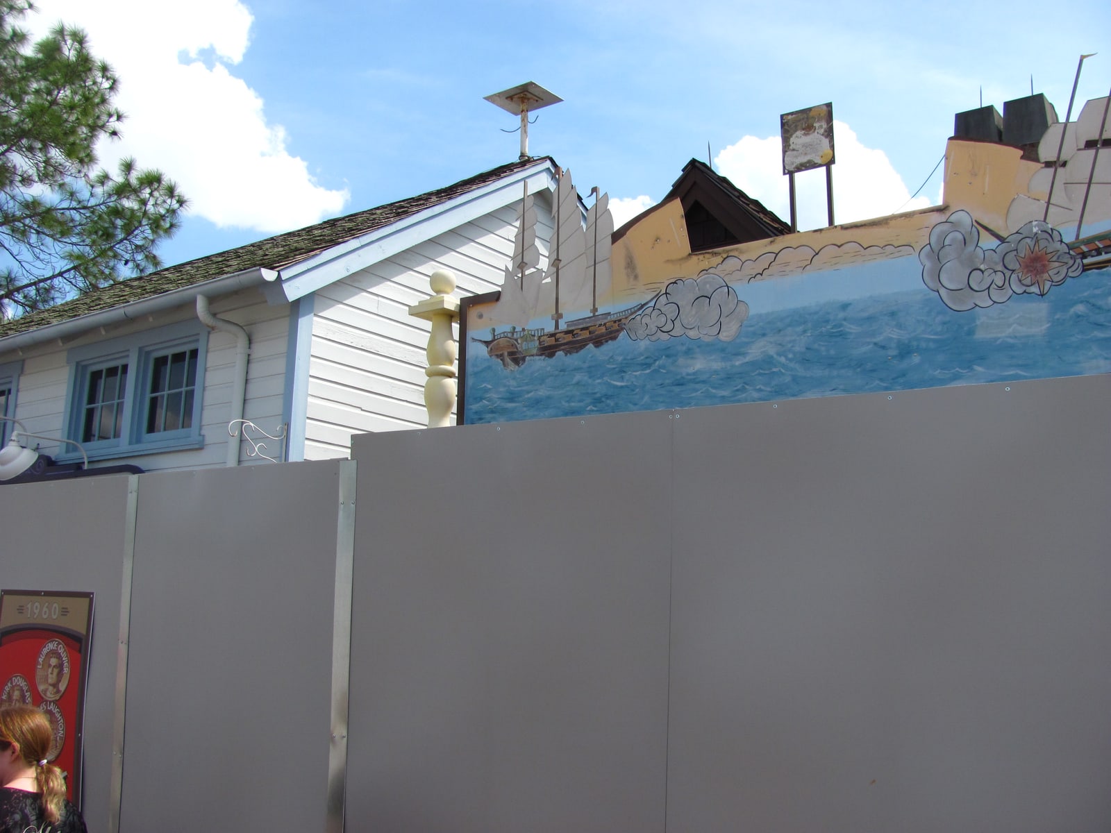 Grey construction walls sealing off a section of the closed Jaws attraction, with a white clapboard Amity building and a painted mural of a tall ship at sea visible above the wall line, and a vintage 1960 movie poster panel at the far left