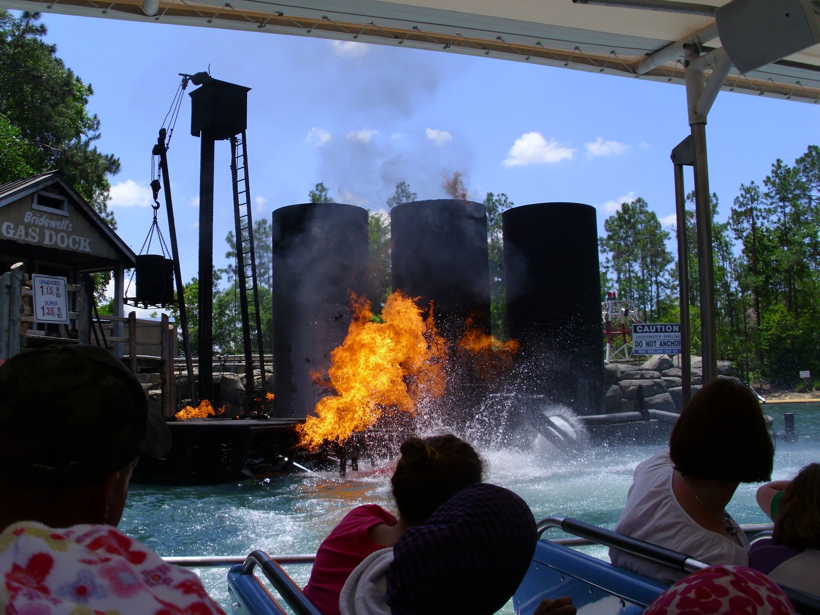 View from a covered tour boat as a wall of orange flame erupts from the water beside a wooden dock marked Bridewell's Gas Dock, with three large black fuel tanks behind it and guests watching from the seats in front