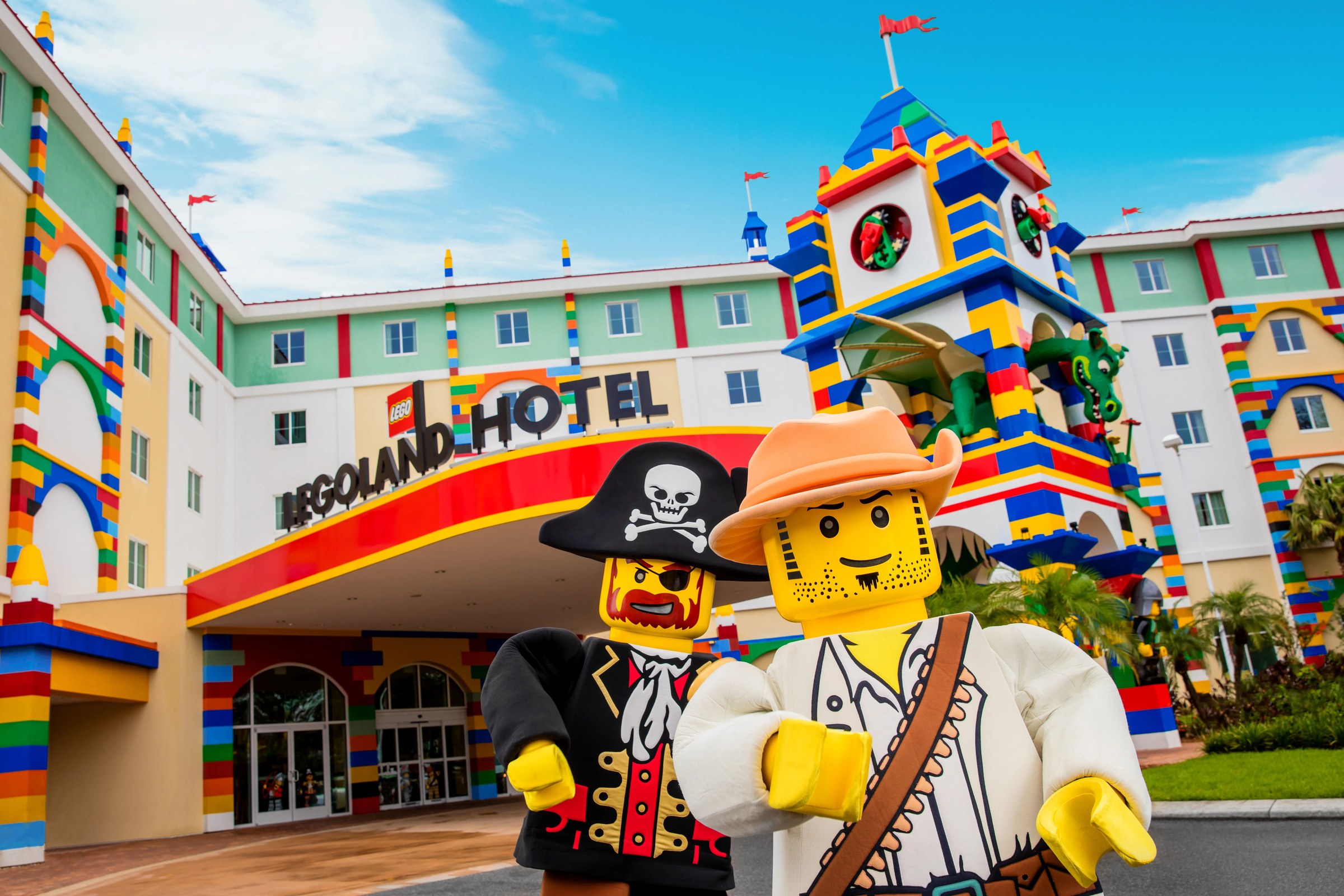 The exterior of the LEGOLAND Hotel on a sunny day, a multistory building in cream, mint green and white trimmed with oversized red, blue and yellow LEGO brick columns. A large sign over the entrance canopy reads LEGOLAND HOTEL, and a tall brick tower with a green LEGO dragon stands at the right. Two walk around characters, a LEGO pirate in a skull and crossbones hat and a LEGO adventurer in a wide-brimmed orange hat, stand in the foreground