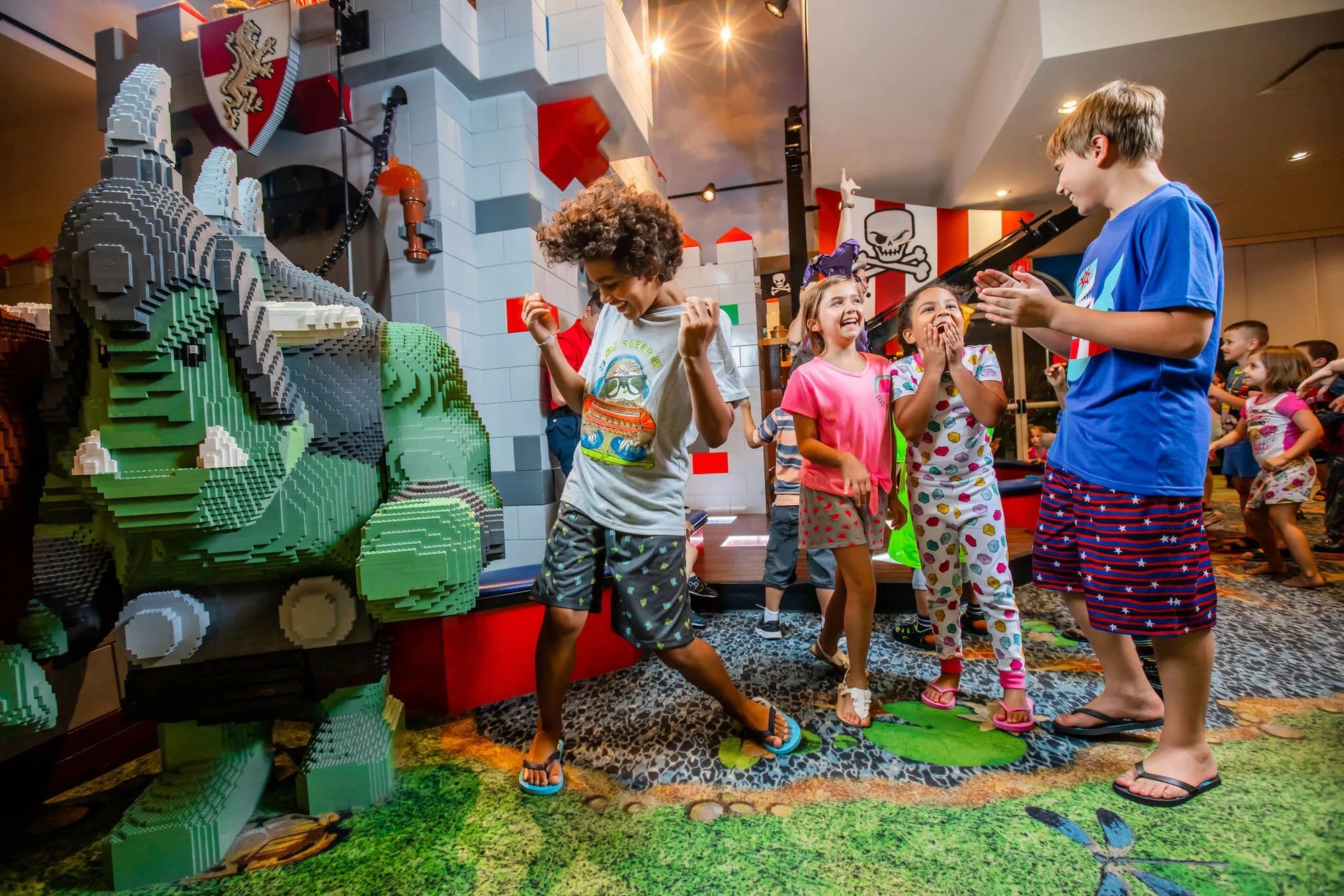 Children, one of them in printed pajamas, dancing on a patterned carpet in a LEGO themed hotel interior. A large green LEGO brick dragon sculpture fills the left of the frame beside a gray LEGO castle wall with a red and white shield bearing a gold lion. Four children in the foreground are laughing and dancing, with more children and adults behind them under warm ceiling lights
