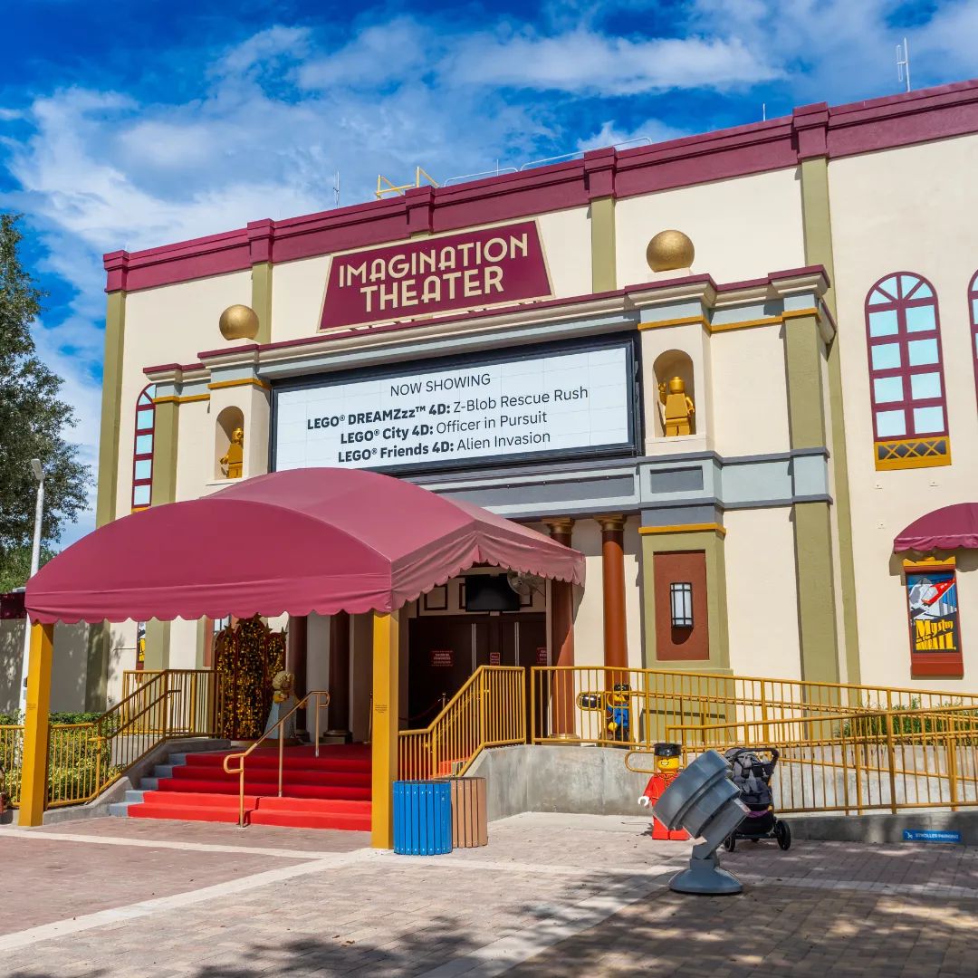 The theater entrance seen from the front left, with the IMAGINATION THEATER sign and NOW SHOWING board above, a maroon dome awning over red carpeted steps, gold handrails, a red LEGO minifigure statue operating a searchlight prop at right and a stroller parking sign behind it