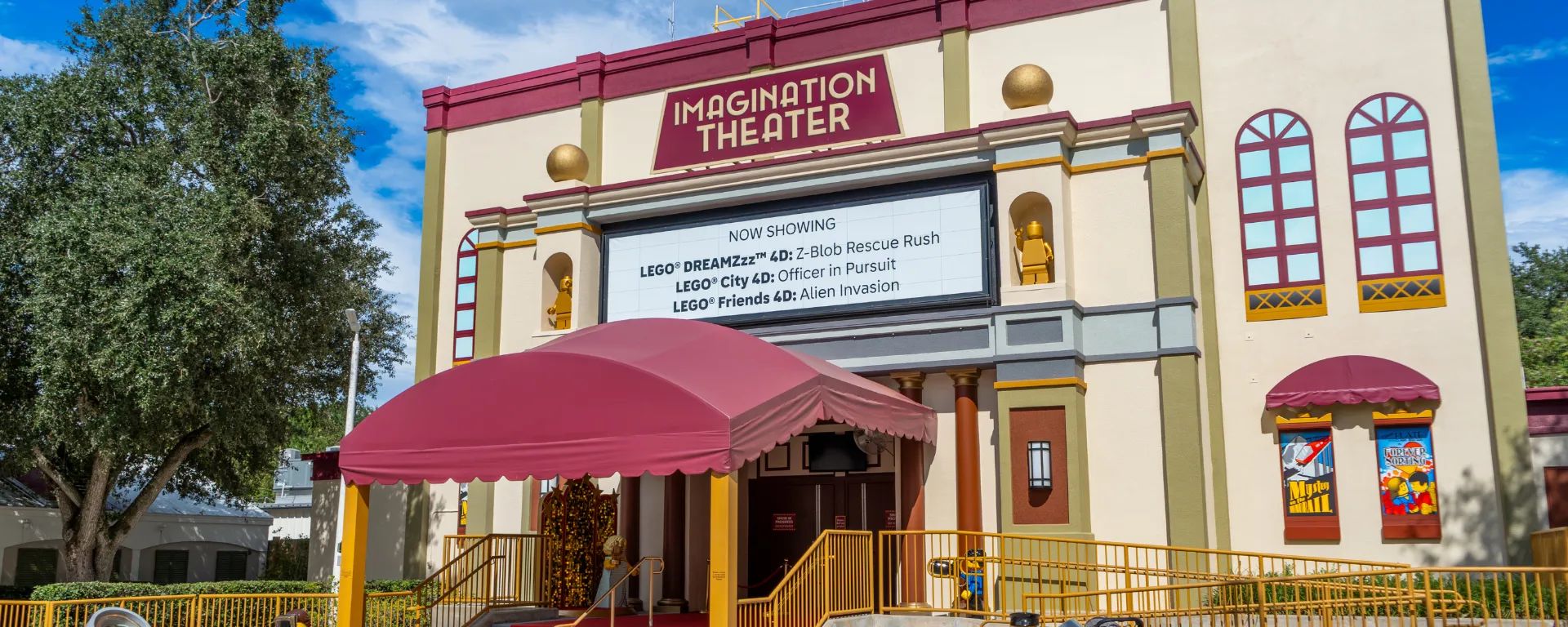 The theater exterior in cream and maroon with gold spheres and arched windows, a maroon sign reading IMAGINATION THEATER above a white NOW SHOWING board listing LEGO DREAMZzz 4D Z-Blob Rescue Rush, LEGO City 4D Officer in Pursuit and LEGO Friends 4D Alien Invasion, with a maroon awning over the entrance and gold minifigure statues in wall niches