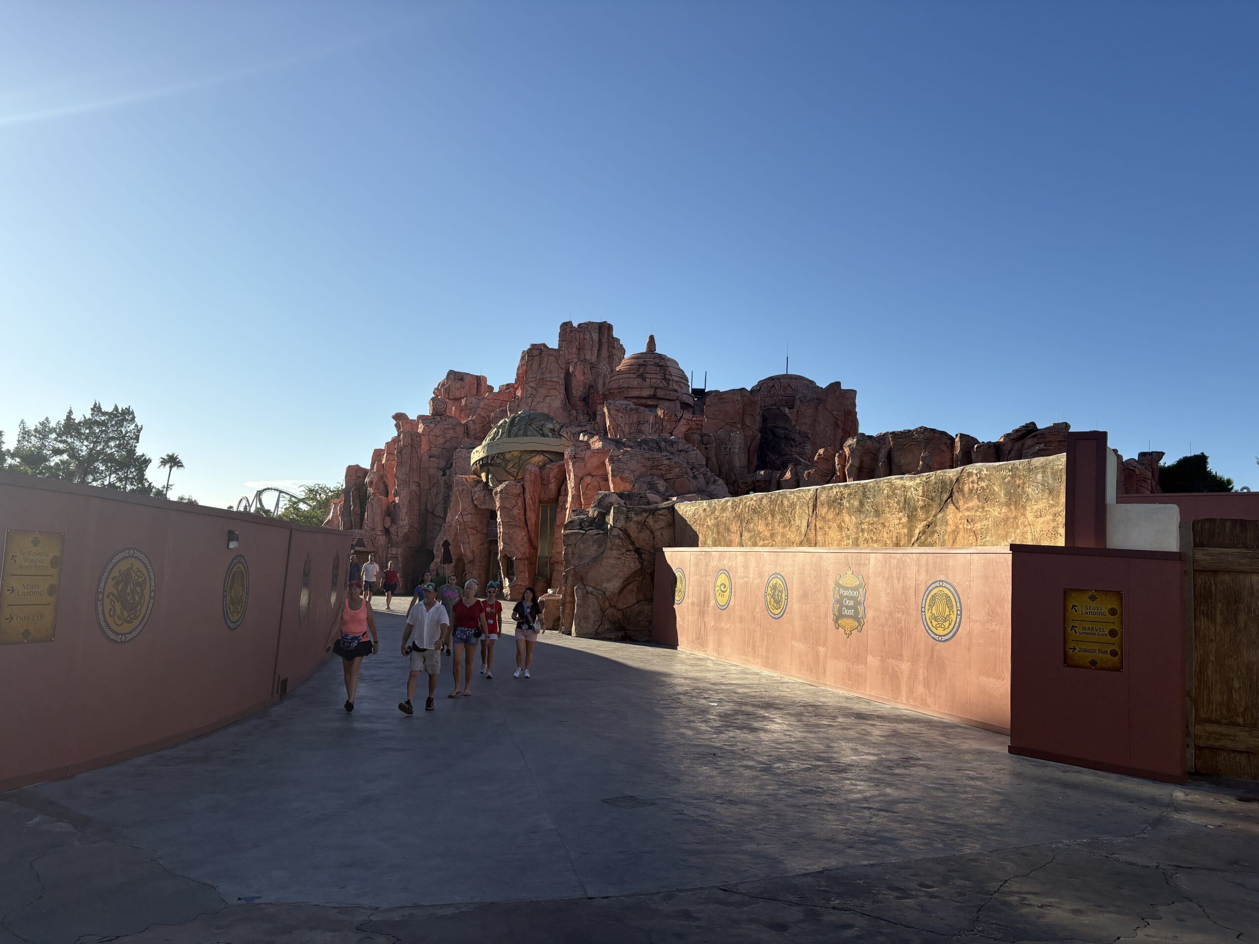 A wide walkway with matching rust orange construction walls running down both sides, each carrying gold medallions and yellow wayfinding signs. Guests walk toward the camera between them. Ahead the path opens onto tall red rock formations and a weathered green and gold dome. A coaster track is visible above the trees at the far left