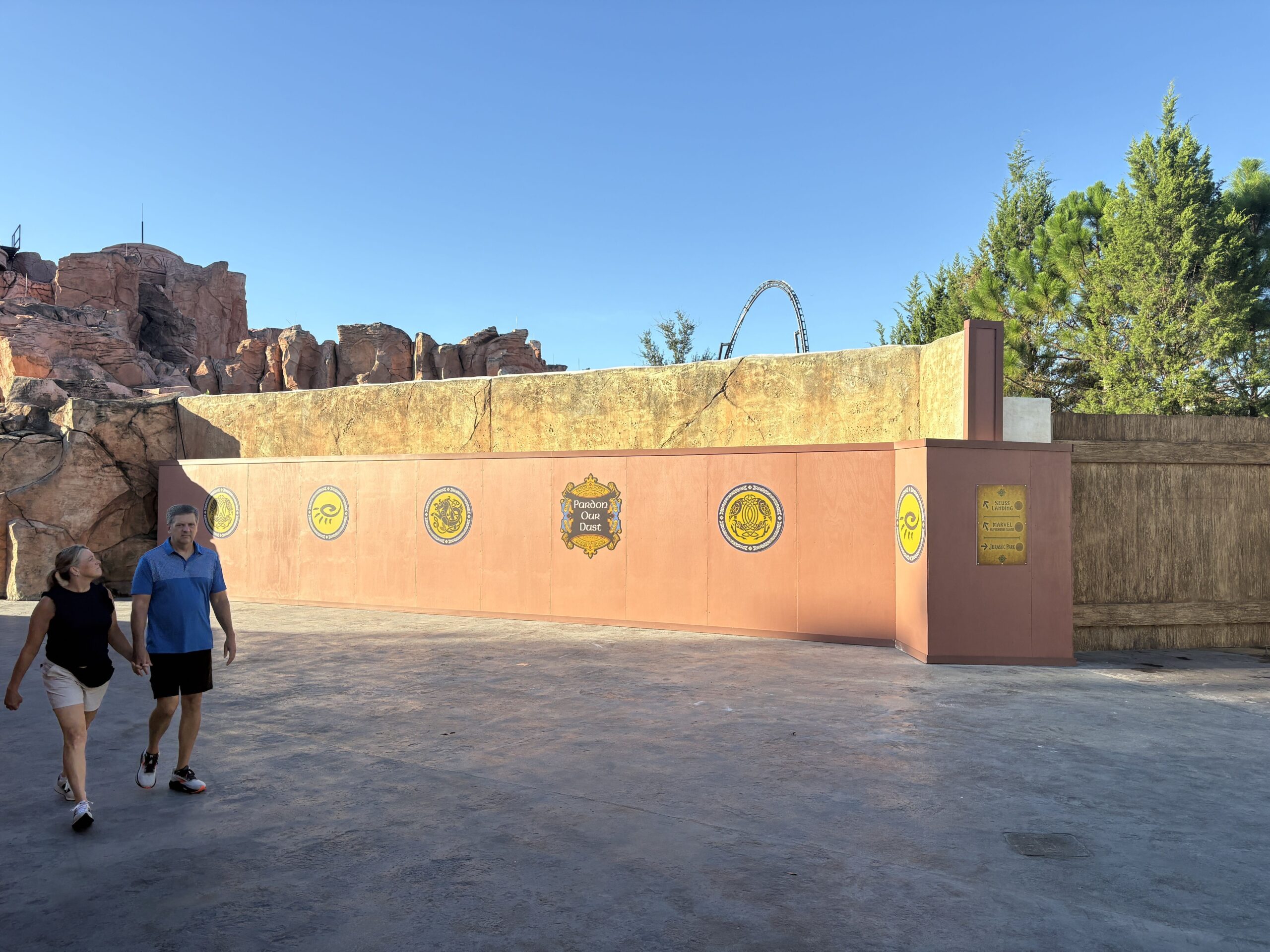 A rust orange construction wall stretching along a wide empty walkway, topped by a tan rock wall. Gold medallions and a decorative Pardon Our Dust plaque are spaced along it, and a yellow wayfinding sign at the right points to Seuss Landing, Marvel Super Hero Island and Jurassic Park. Red rock formations rise at the left and a steel coaster loop is visible in the distance. A couple walk past in the foreground