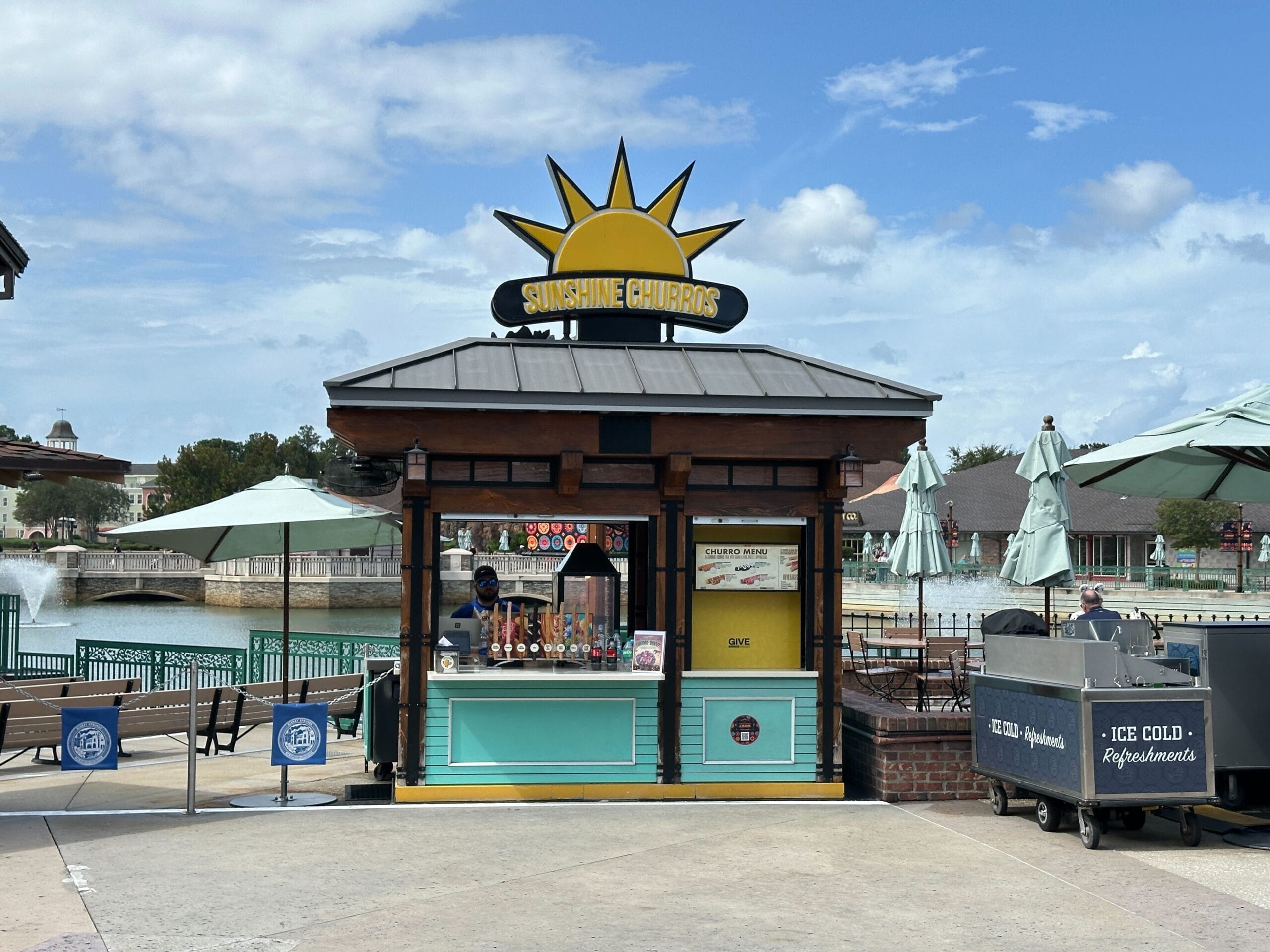 A Sunshine Churros kiosk open for business at Disney Springs, with a yellow half-sun sign above a black oval reading Sunshine Churros on the peaked metal roof, a cast member behind the turquoise counter beside a lit glass warmer holding upright churros, and a Churro Menu board on a yellow panel to the right