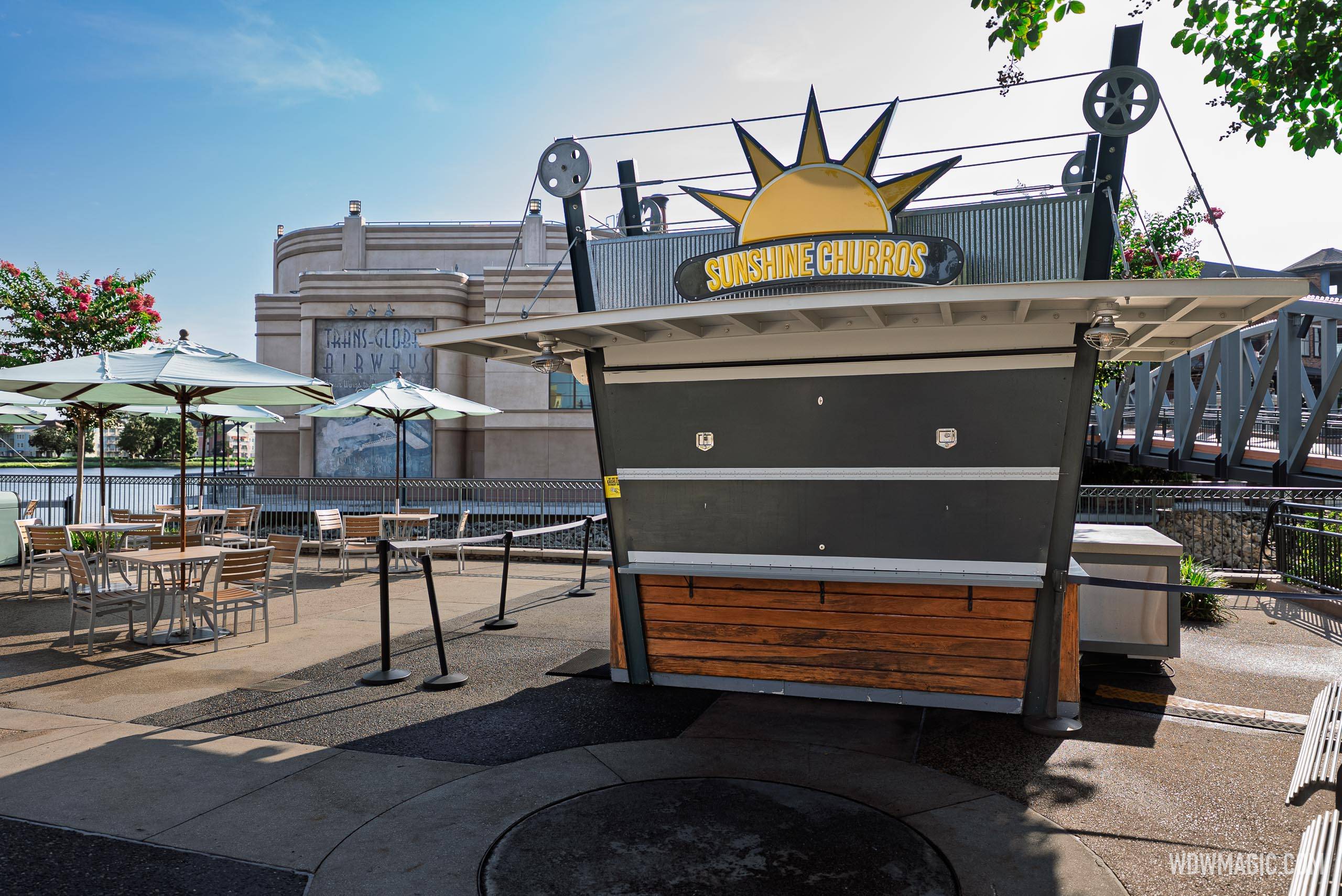 The West Side Sunshine Churros kiosk with its service window closed behind a flat gray panel, no staff and no product visible, under an angled white canopy on black posts with film reel shaped pulleys, empty queue stanchions in front and the Art Deco Trans-Global Airways facade behind