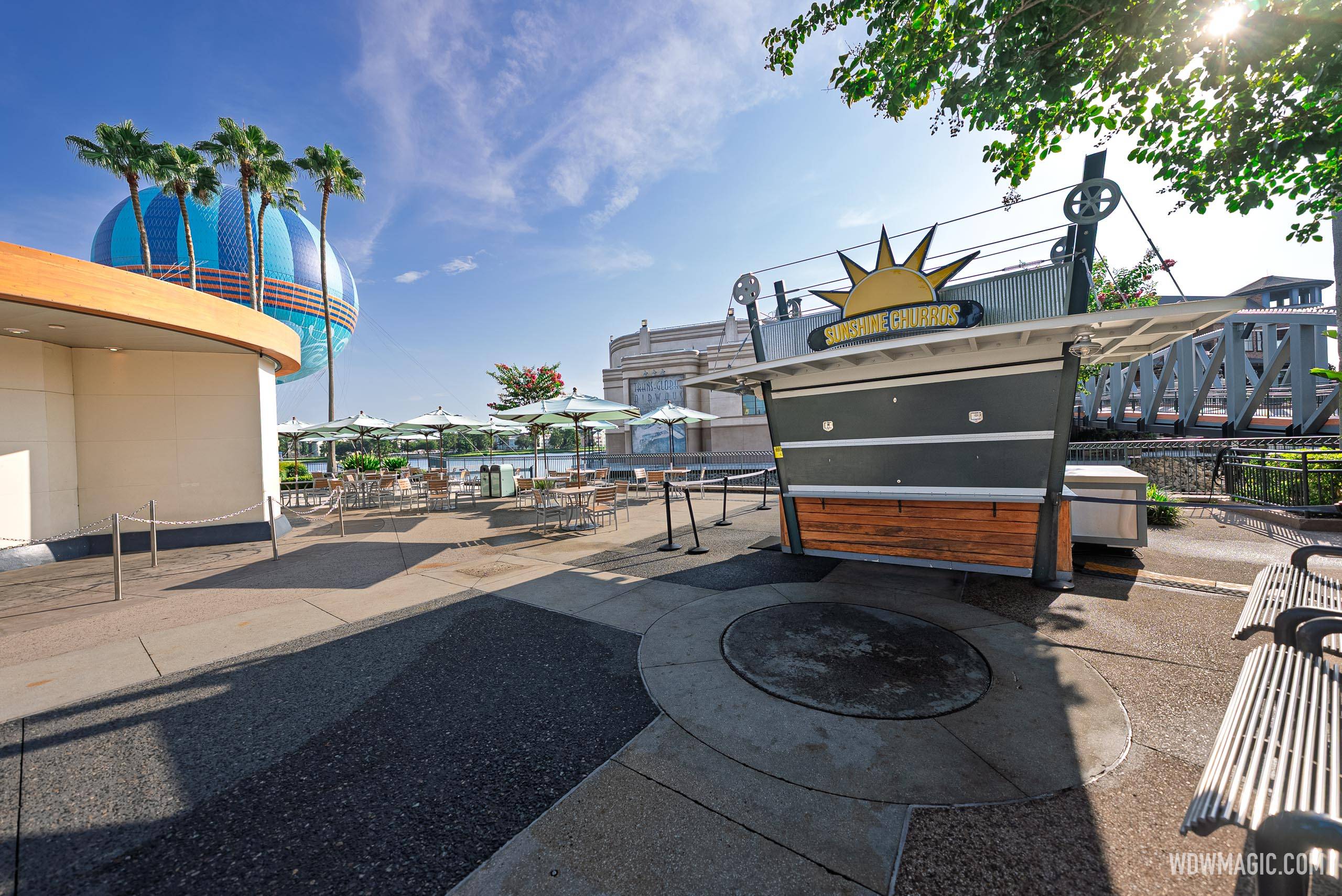 A wider view of the shuttered West Side Sunshine Churros kiosk from further left, with empty umbrella tables, tall palms, the blue Aerophile balloon with its orange stripe inflated behind the palms, and a gray steel truss bridge at right