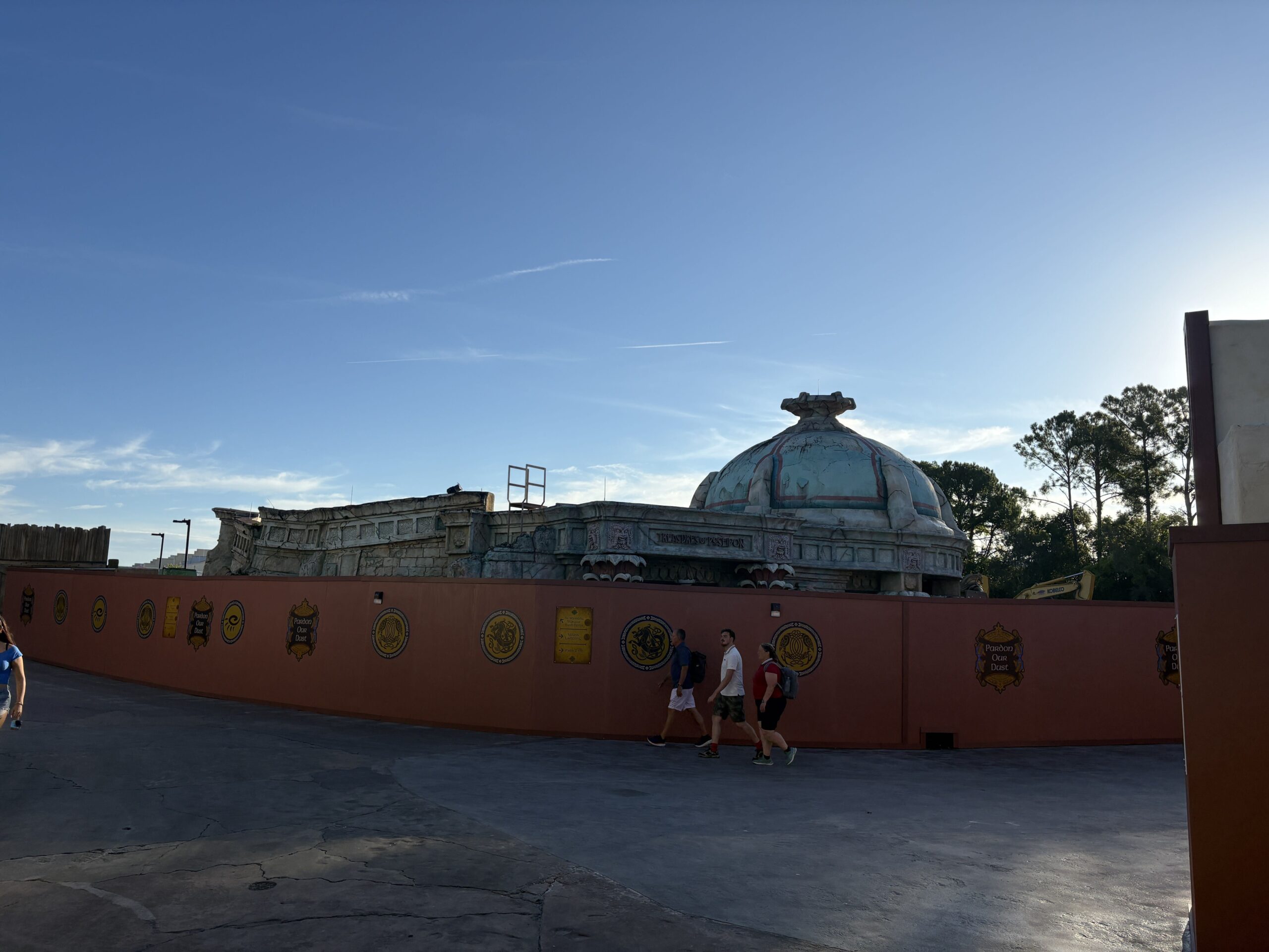 A long rust orange construction wall runs across the frame, decorated with gold and blue medallions and several Pardon Our Dust plaques. Rising behind it is a weathered stone building with a domed roof in faded teal and rust, and a carved sign on its frieze reading Treasures of Poseidon. A yellow excavator is parked behind the wall at the right. Guests walk along the path in front of the wall under a blue sky