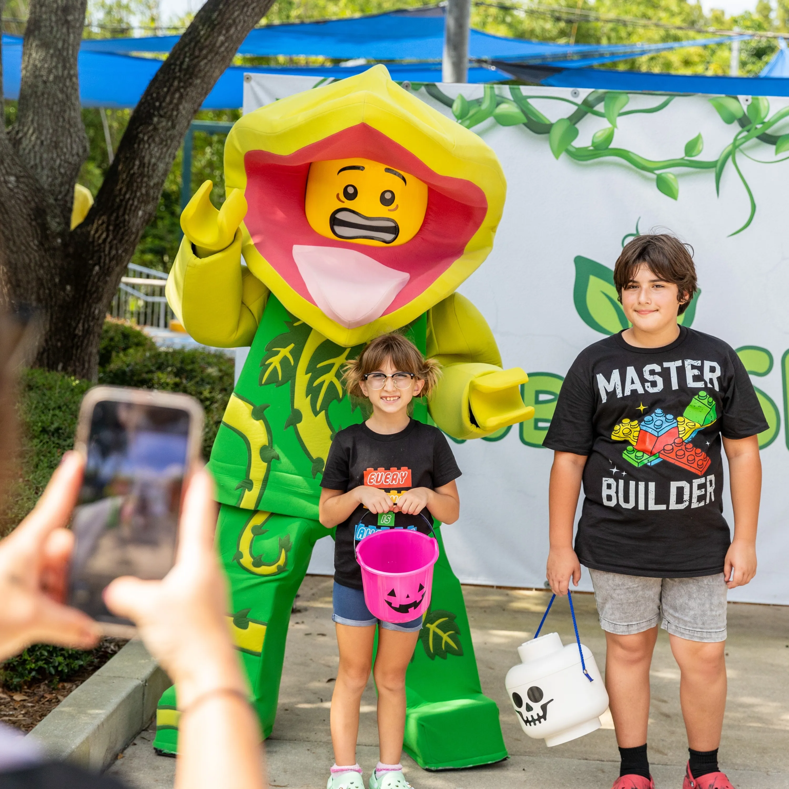 The Plant Monster walkaround character, a green figure with a yellow and pink flower head, posing outdoors in daylight with two children who are holding a pink jack-o-lantern bucket and a white LEGO skull bucket, while an adult photographs them with a phone.