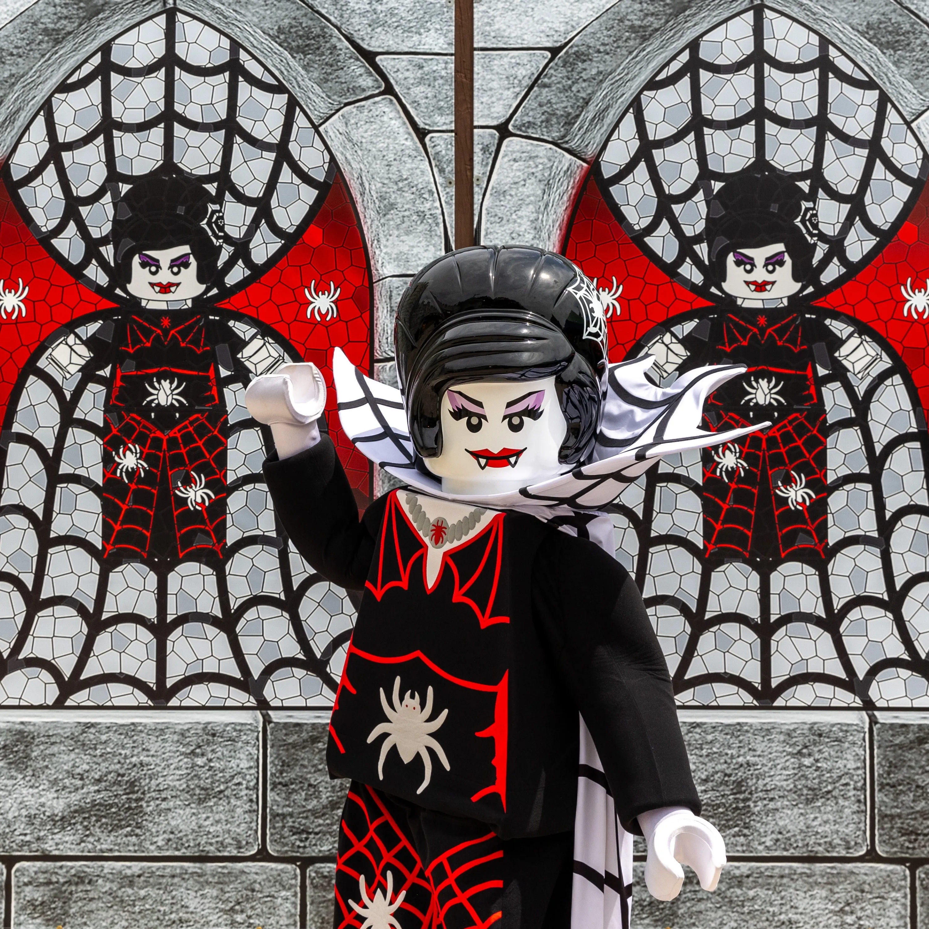 The Spider Lady walkaround character in a black and red spiderweb dress with a tall black hairpiece, posing with one arm raised in front of a grey castle wall with two arched stained-glass windows showing her own likeness on red and white webbing.