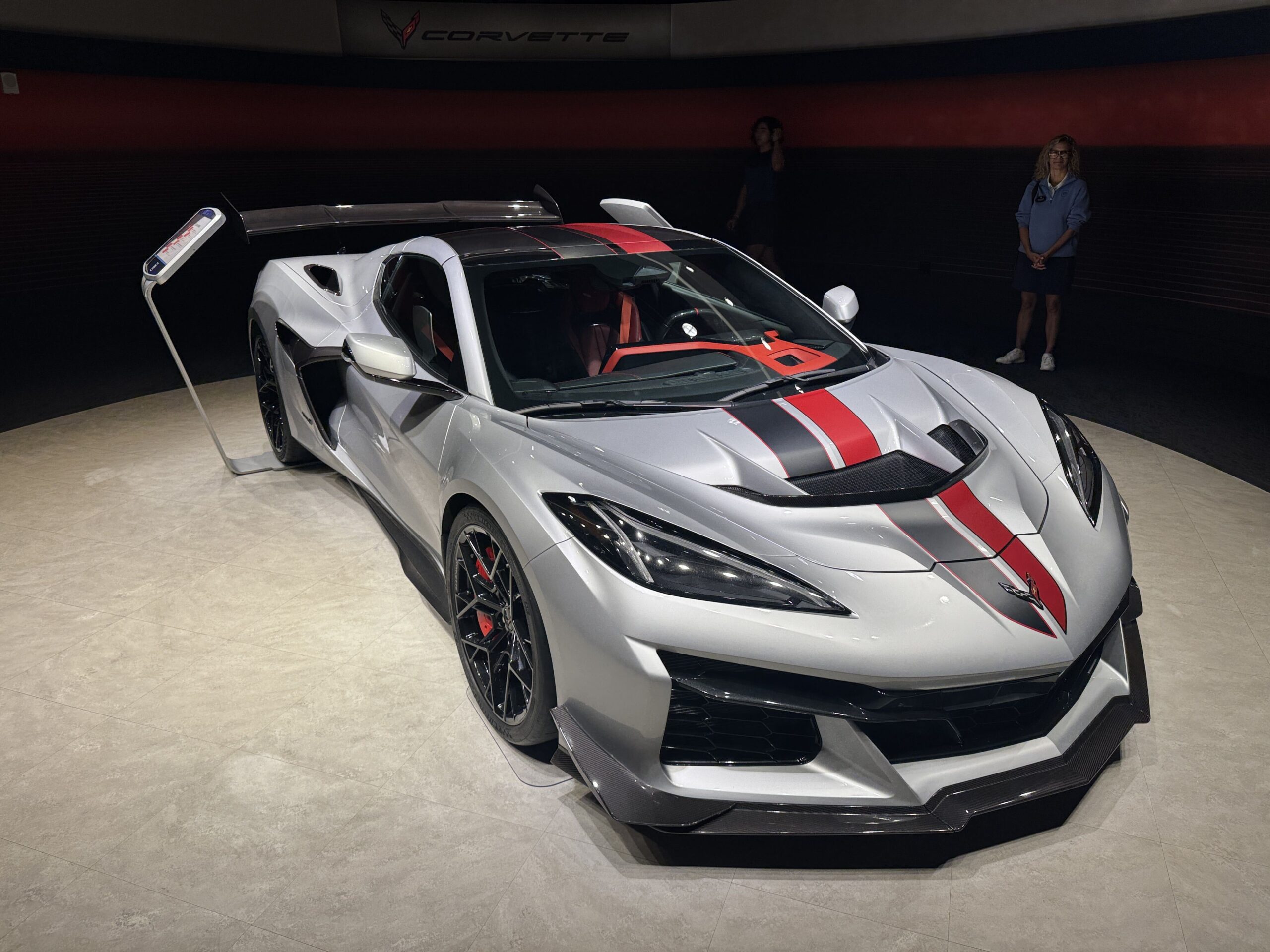 The Corvette seen from the front on the circular showroom floor, with a lit Corvette wordmark sign on the curved wall above and two guests standing in the darkened showroom behind.