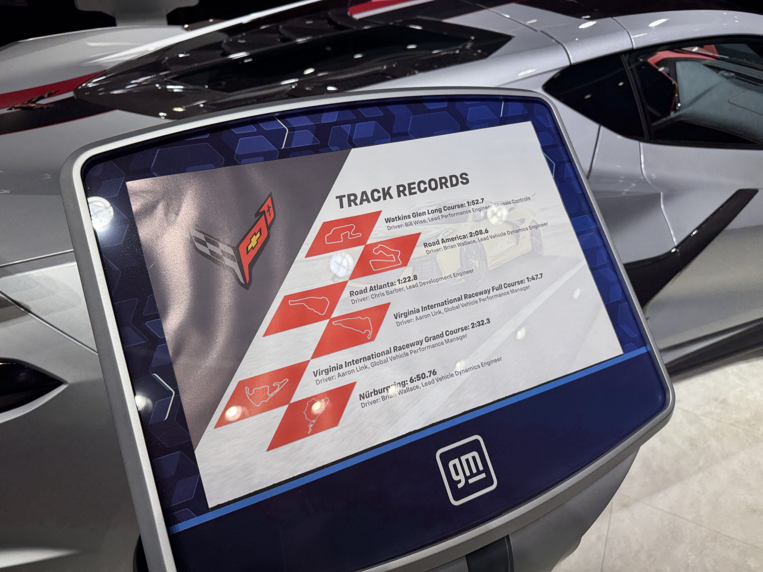 The reverse of the display placard, headed TRACK RECORDS, listing six lap times with track outline diagrams and driver names, with a GM logo at the bottom and the silver Corvette behind it.