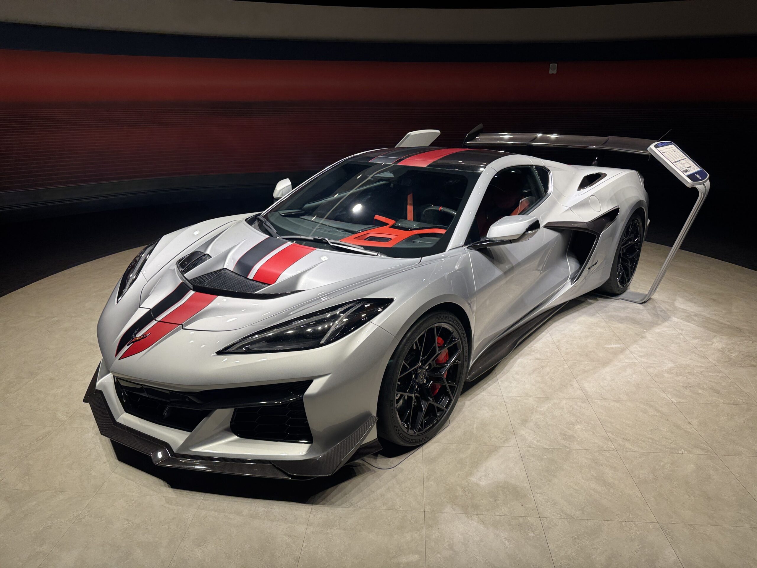 A silver Chevrolet Corvette ZR1 with red and black racing stripes standing on a tiled circular display area, with a window sticker on a tall stand beside it.