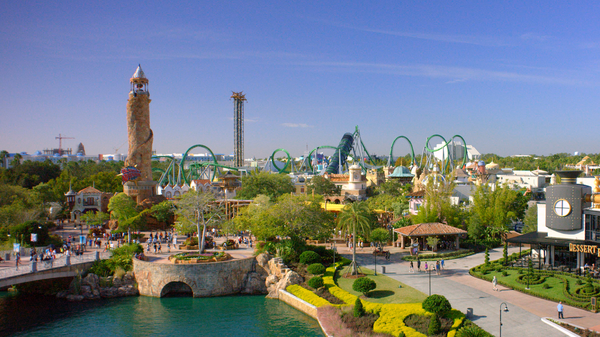 An elevated view toward the entrance of Islands of Adventure on a clear day, with the stone Pharos lighthouse and its Islands of Adventure sign at the left, green Incredible Hulk coaster track and a pair of tall latticed tower ride structures on the skyline, two construction cranes far off at the left horizon, guests crossing the entrance plaza, and the lagoon and a CityWalk restaurant terrace in the foreground.