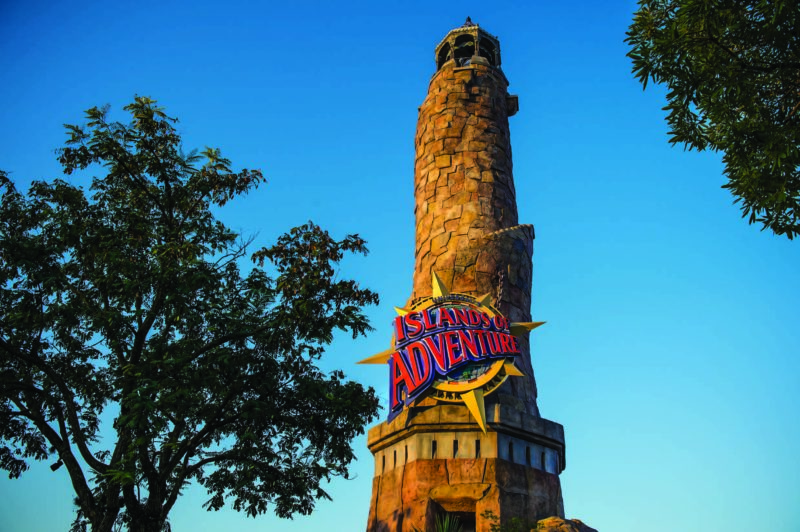 The stone Pharos lighthouse at the entrance to Islands of Adventure photographed from below against a deep blue sky, with the starburst Islands of Adventure sign mounted on its side and tree branches framing the shot.