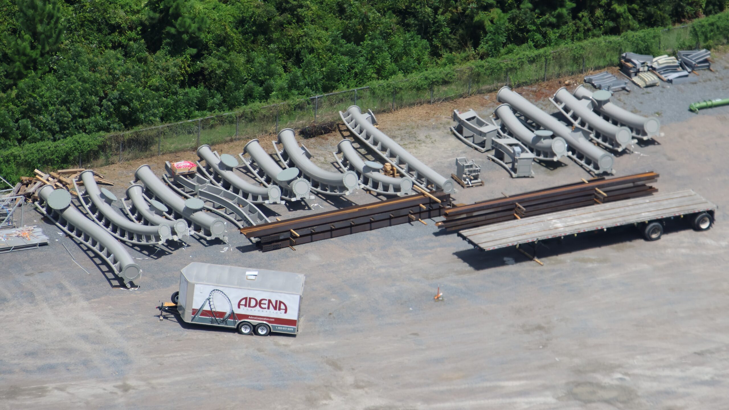 Rows of curved gray coaster track sections resting on cradles in a dirt lot, with a white trailer marked Adena Corporation parked beside them and a flatbed trailer to the right.