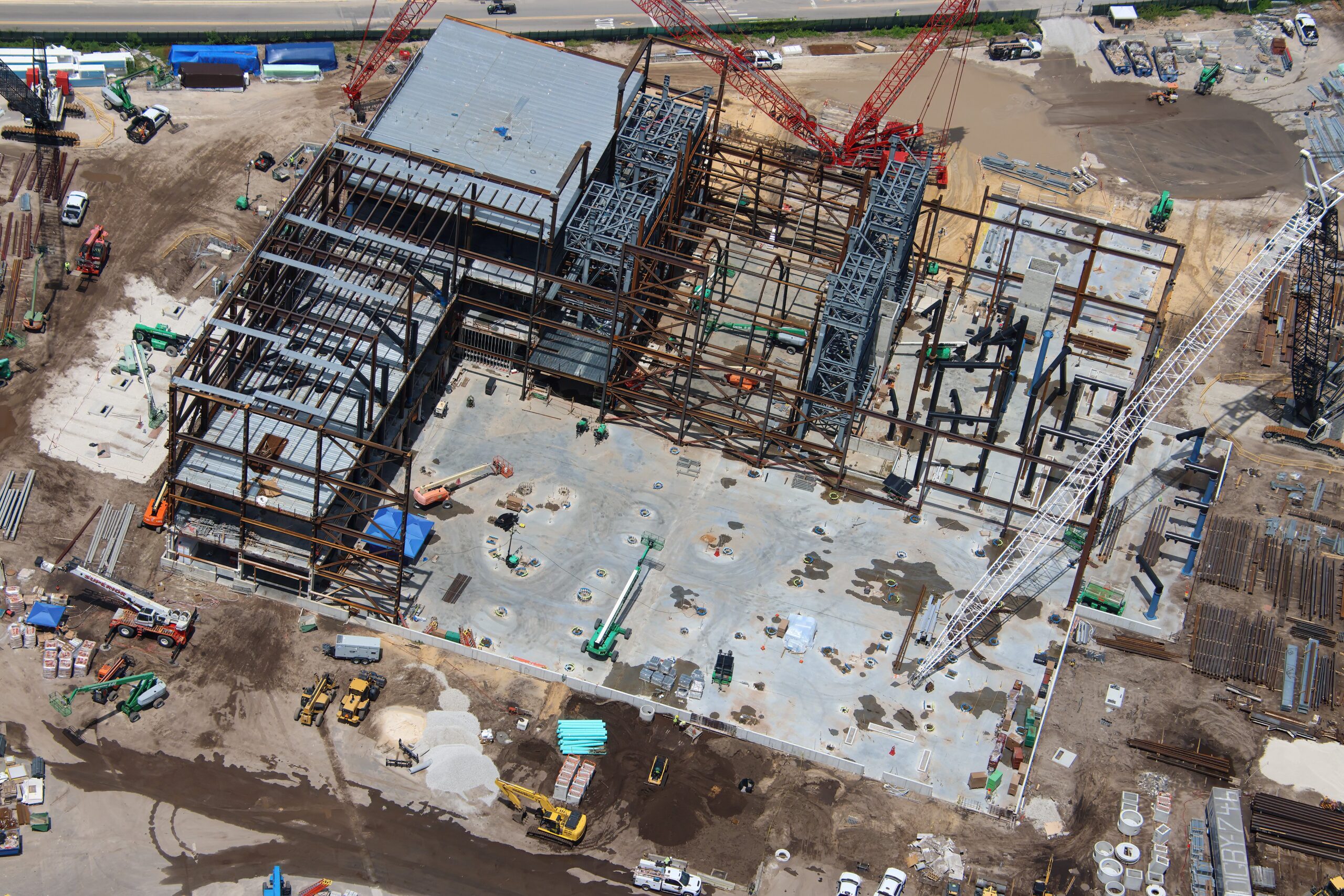 Aerial view of the Monsters, Inc. door coaster show building under construction, showing rust colored structural steel, two gray lattice towers, a partially roofed section and multiple cranes.