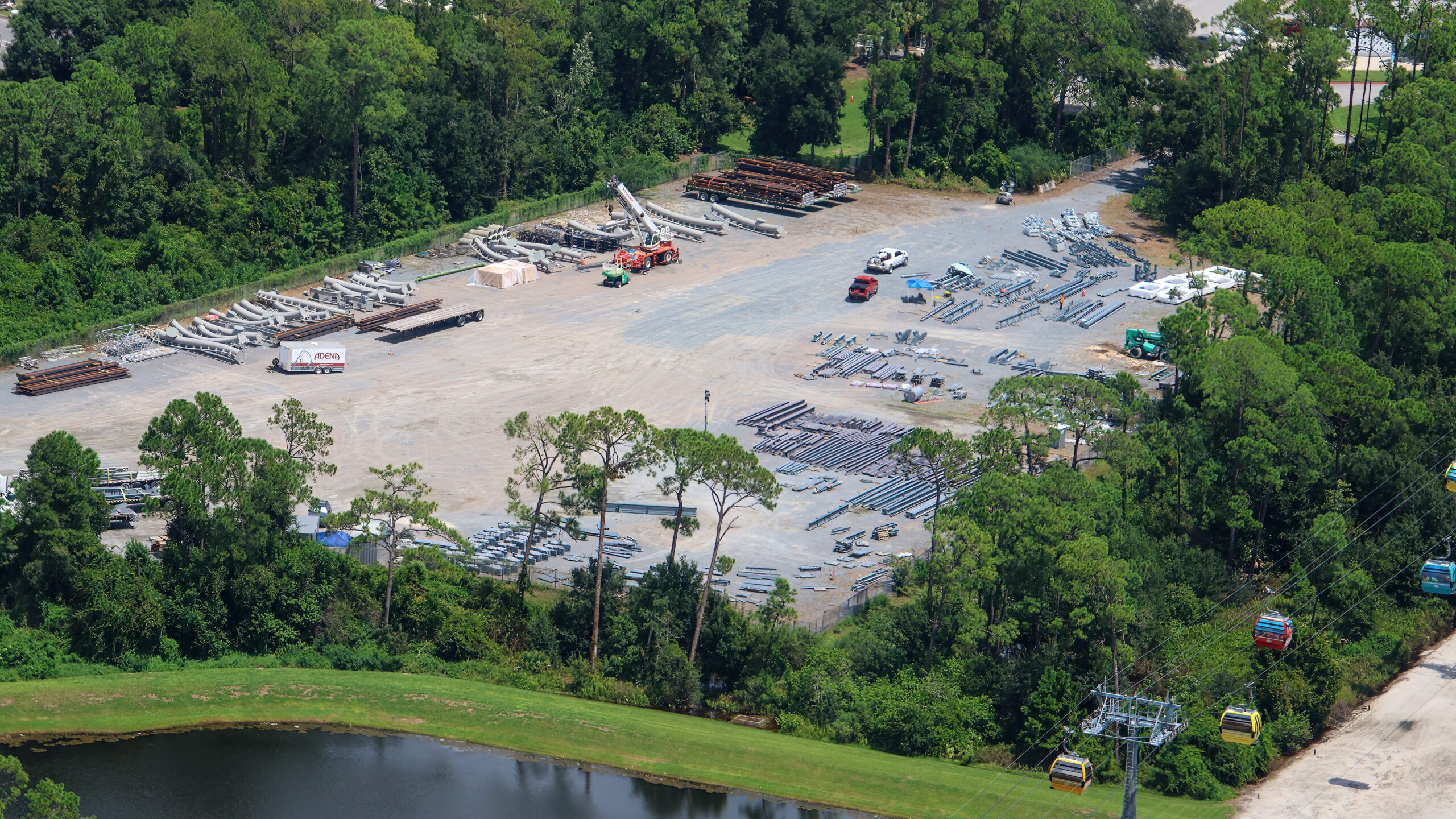 Aerial of a cleared staging yard ringed by trees, holding rows of gray track pieces and steel, with Disney Skyliner gondolas passing on cables at lower right.