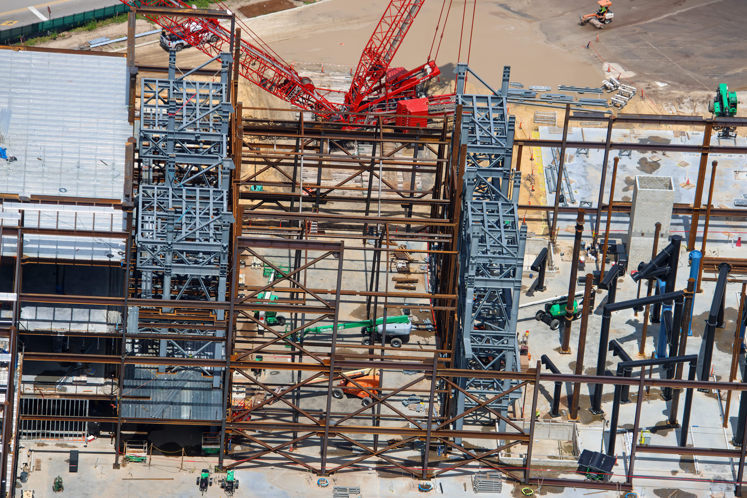 Close aerial of two gray steel lattice towers standing on either side of rust colored structural steel bays, with a red crane working overhead.