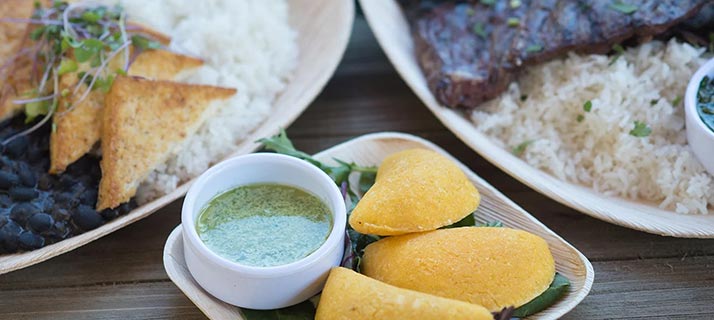 Food at Waterway Grill: yellow empanadas on a leaf lined dish beside a small white cup of green sauce, with plates of white rice, black beans, fried triangles and glazed ribs around them on a wooden table.