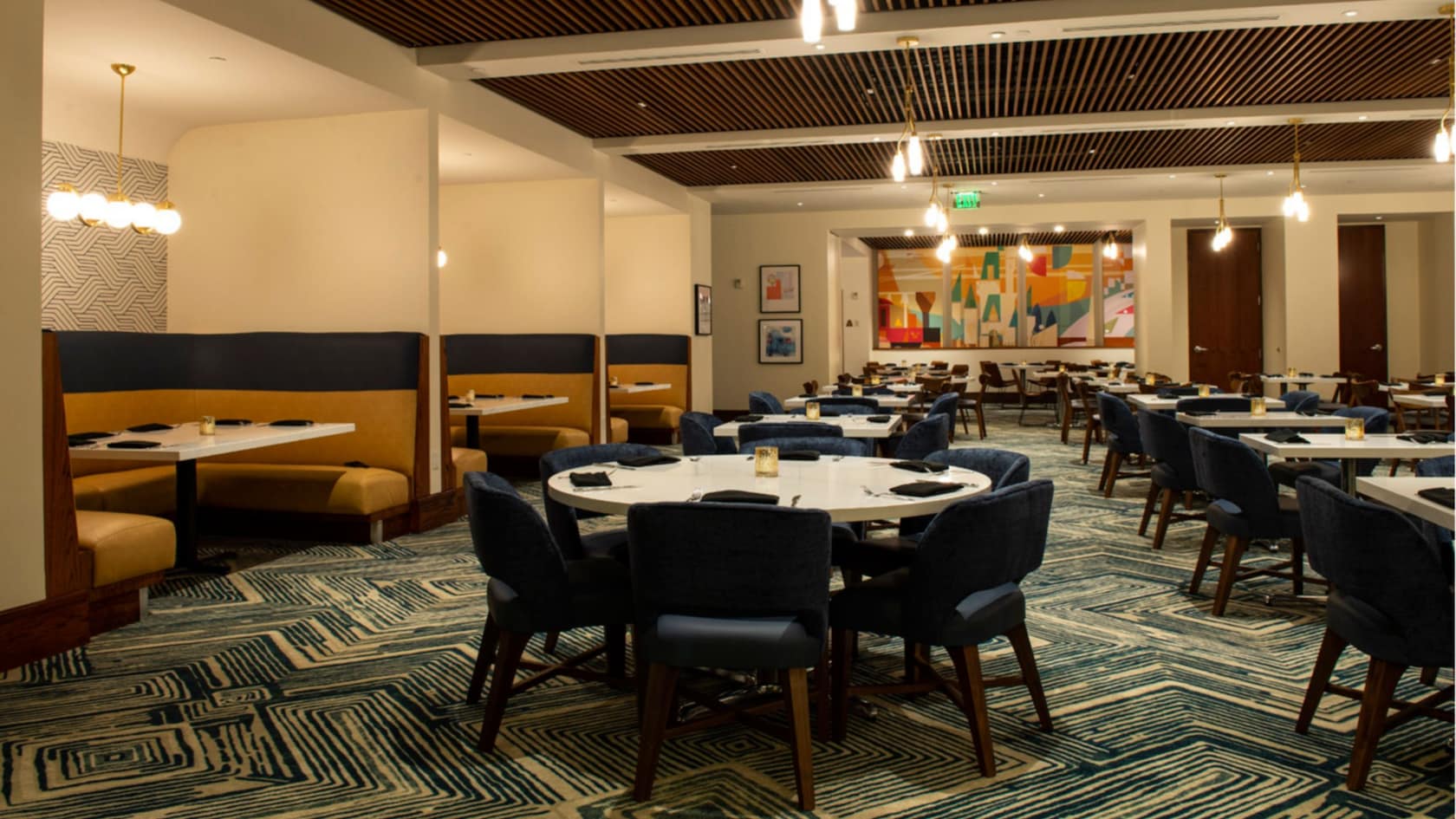 The empty Steakhouse 71 dining room, with mustard yellow and navy banquettes along the left wall, round white marble topped tables with navy tub chairs, a slatted wood ceiling with brass pendant lights, and a large colorful mural on the far wall.