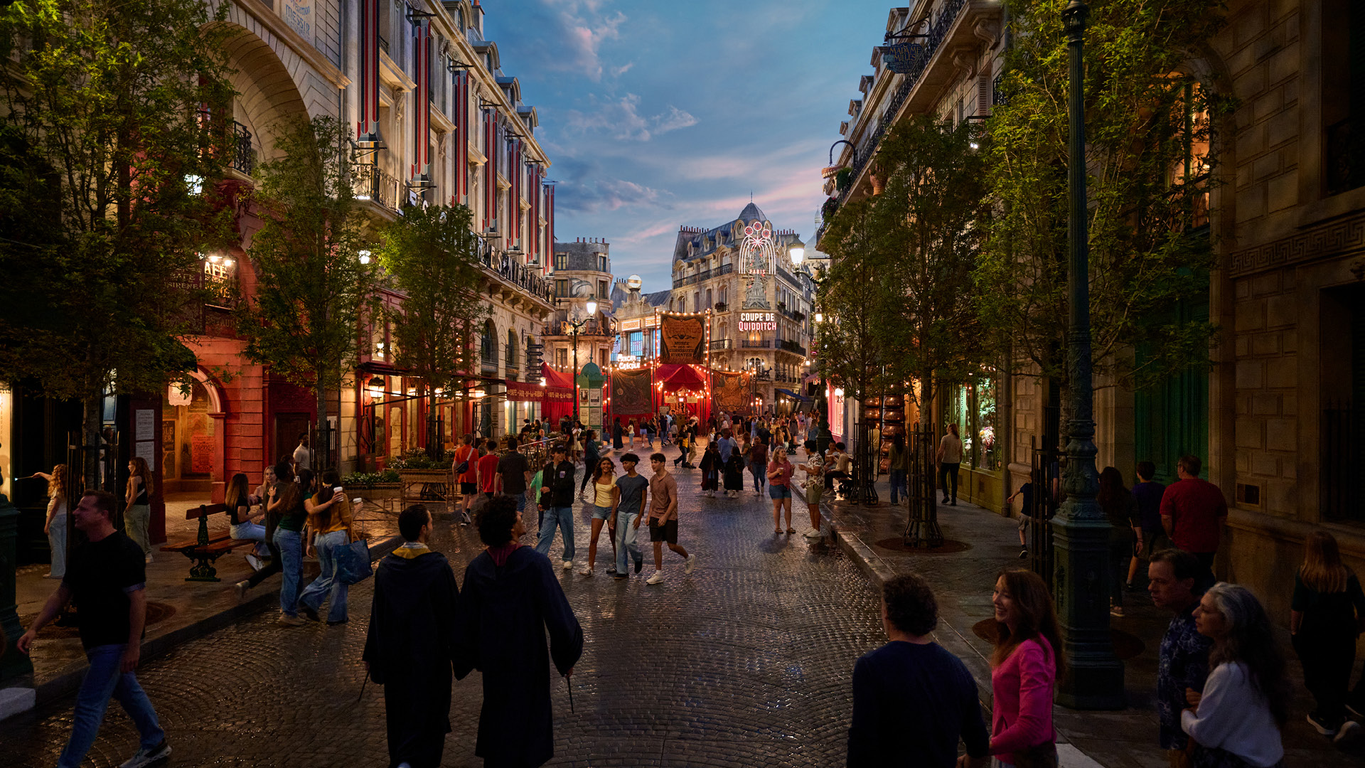 Guests walking down a Paris street at Epic Universe at dusk, two of them in long flowing wizard robes in the foreground, with hanging fabric banners strung across the street ahead.
