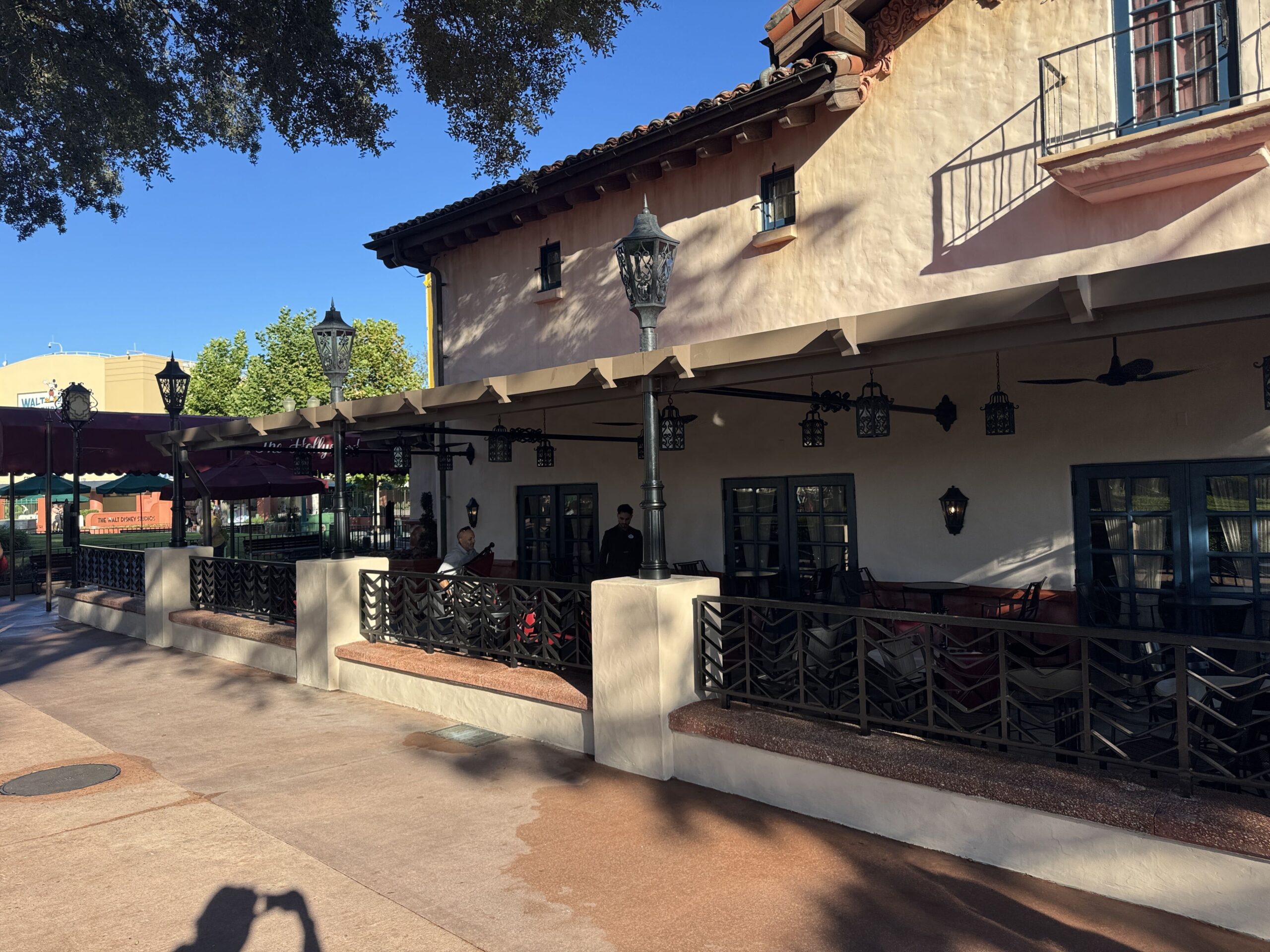 The exterior of The Hollywood Brown Derby at Disney's Hollywood Studios, with a new overhang running along the side of the building. Black lanterns and ceiling fans hang beneath it, black lampposts rise from white stucco column bases along a black chevron patterned railing, and the older maroon awning and a matching umbrella are visible further down the walkway.