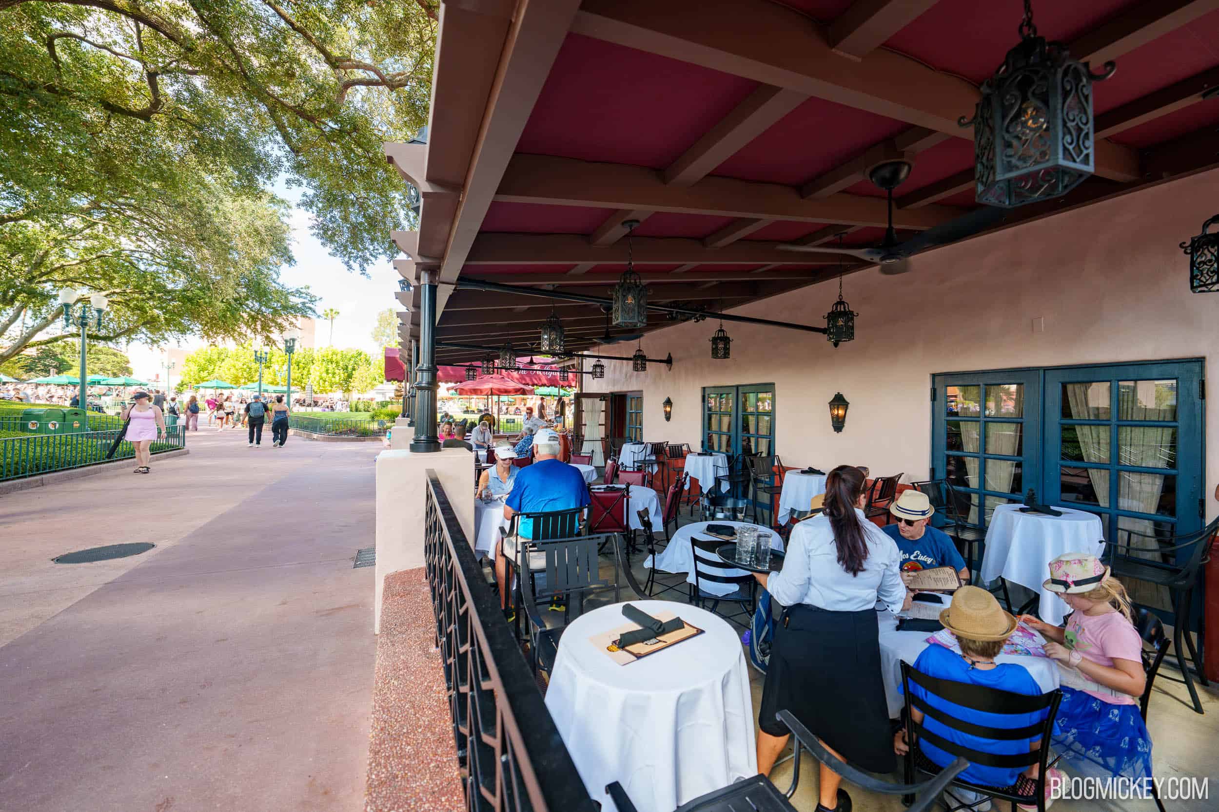 Guests dining on the covered Brown Derby Lounge patio. Deep red painted beams run across the underside of the new overhang, with wrought iron style lanterns hanging on chains and a black ceiling fan. Round tables with white tablecloths are set among black metal and red leather chairs, a server attends a table, and the park walkway runs alongside.