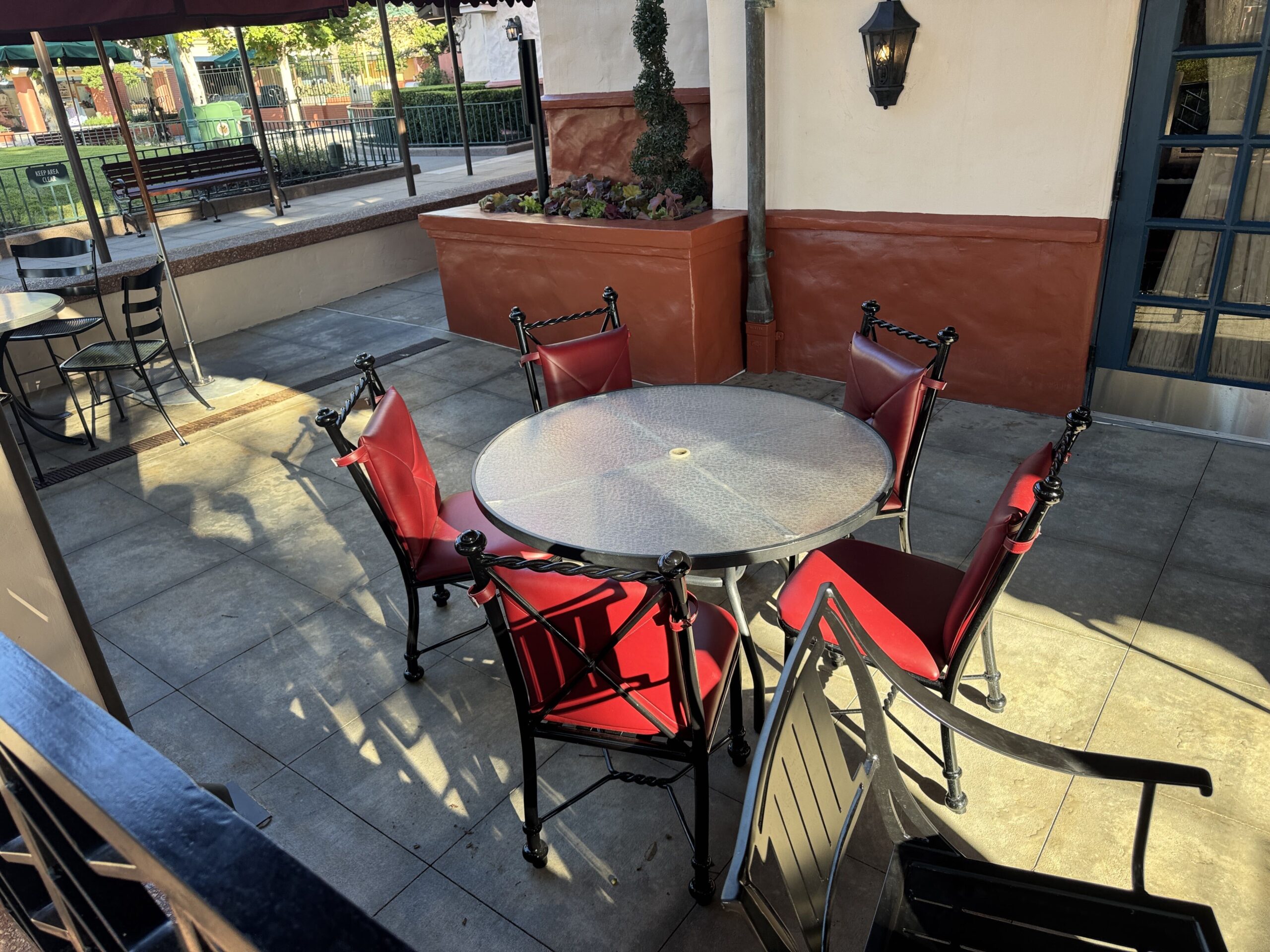 A round glass topped table on the Brown Derby Lounge patio surrounded by chairs with bright red upholstered seats and backs on black wrought iron frames with twisted rope detailing, standing on newly laid pavement beside a terracotta planter.