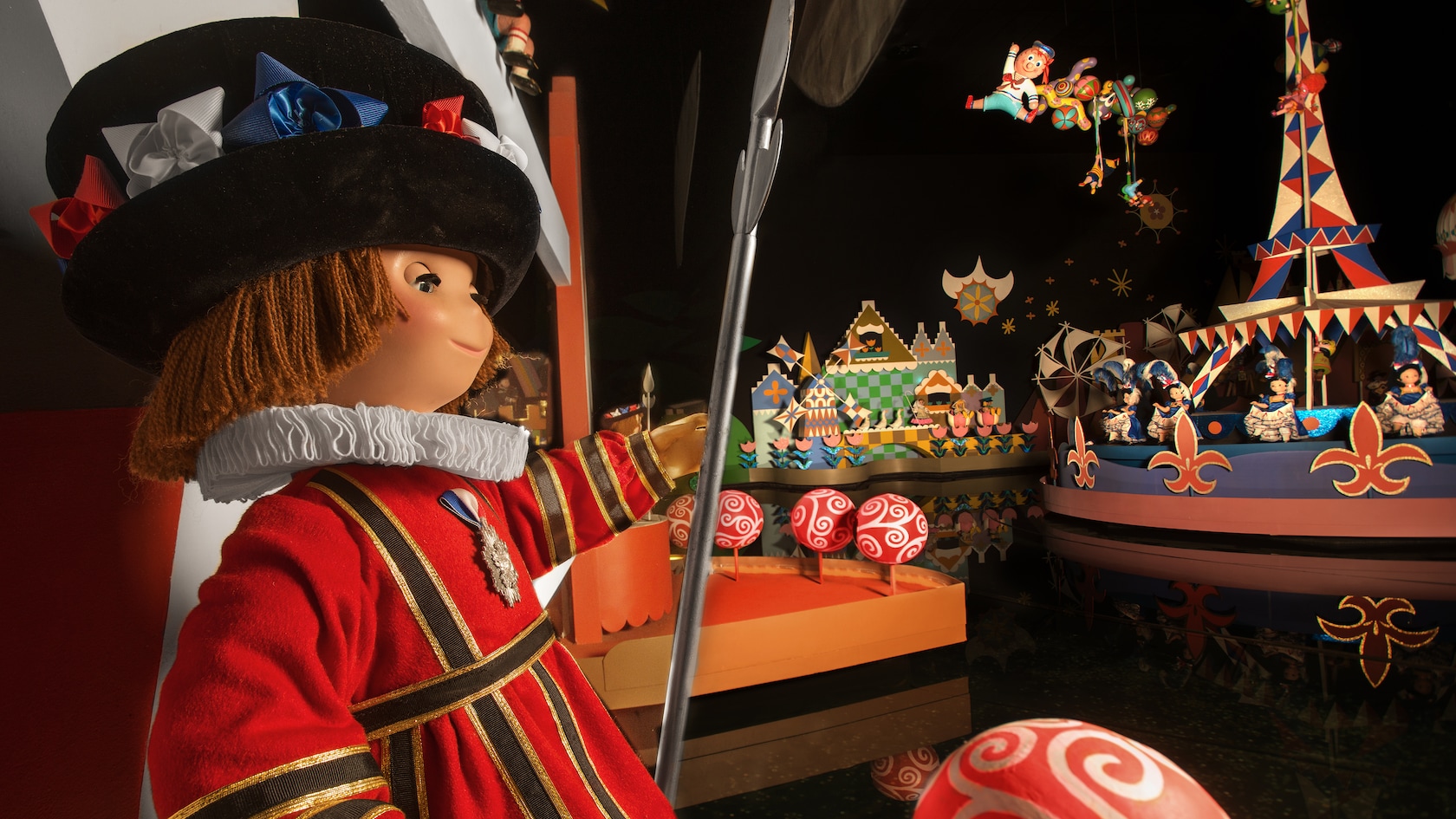A Beefeater doll in a red and gold tunic with a white ruff collar and a wide black hat trimmed with red, white and blue ribbon rosettes holds a tall silver spear inside the Europe room of it's a small world. Behind him are a red, white and blue Eiffel Tower hung with pennants, dolls in blue and white dresses standing on a fleur de lis boat, red and white swirled lollipop props, and a doll suspended from a cluster of balloons overhead.