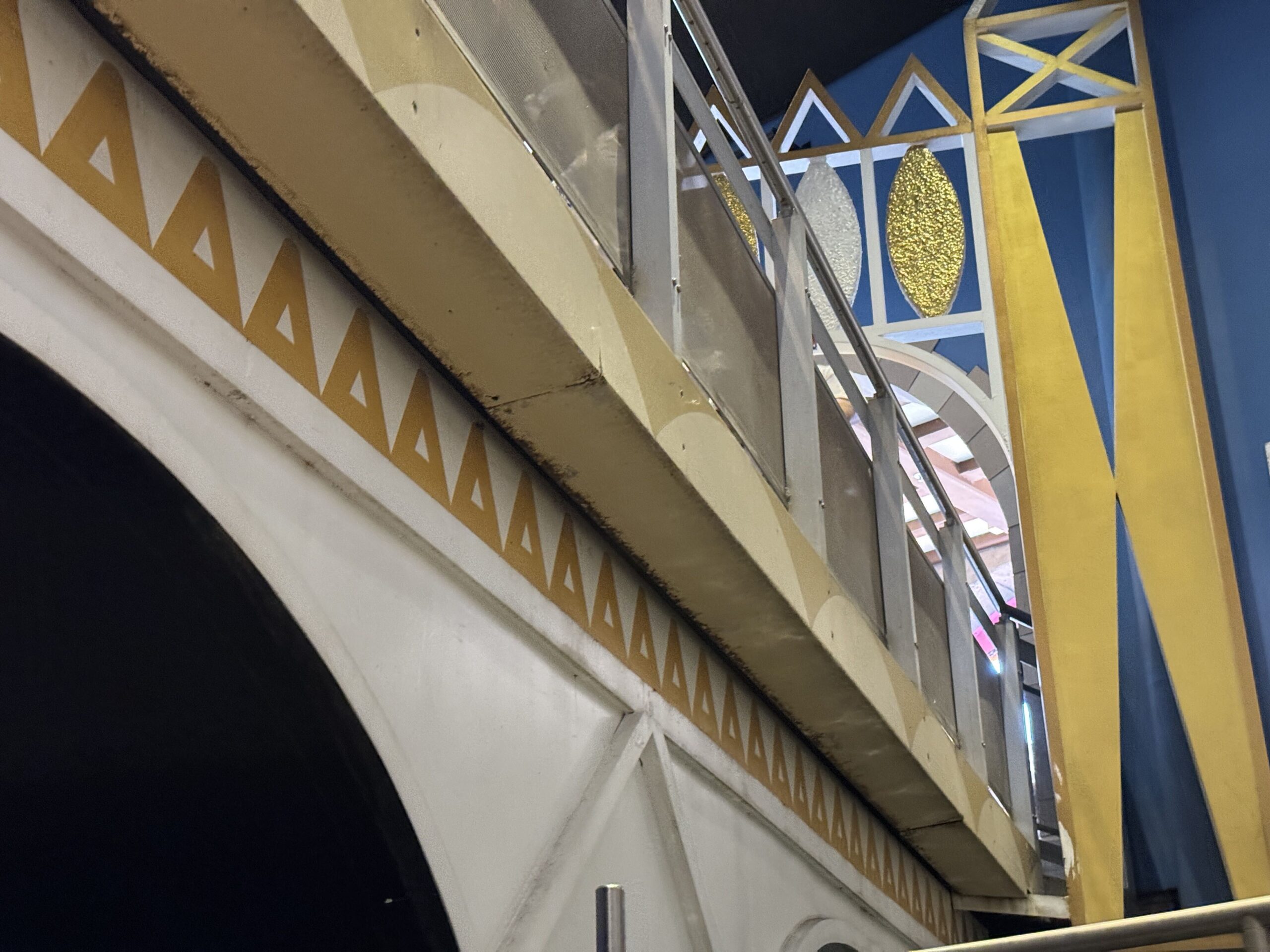 Looking up at the underside of the queue bridge inside it's a small world. A white arch band decorated with a row of gold triangles runs diagonally across the frame, and a dark grimy line runs along the underside edge of the gold-colored beam above it. Gold and white lattice railing is visible at upper right against a deep blue backdrop.