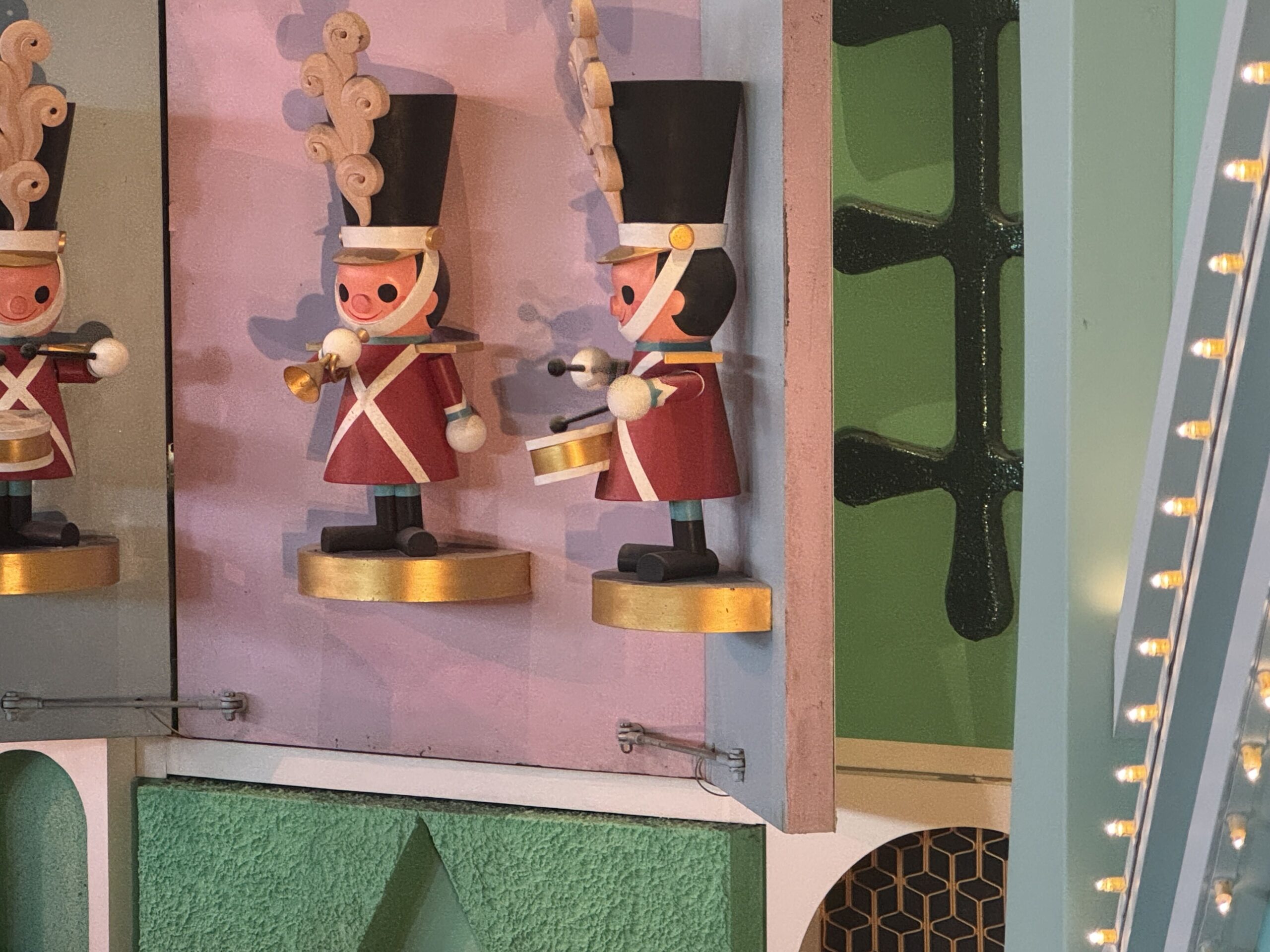 Three wooden toy soldier figures on the it's a small world facade, in red tunics with white cross straps and tall black hats topped with pale curled plumes, standing on gold half round plinths against a pink panel. One holds a gold trumpet and one holds a gold drum with drumsticks. Thin metal rods and a wire run along the bottom edge of the panel, with a bulb lit teal arch at right.