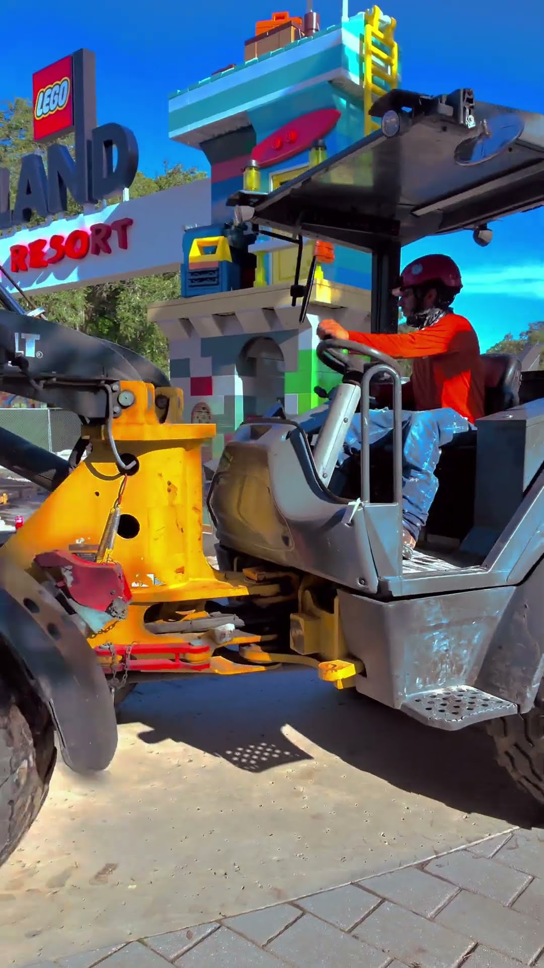A worker in an orange long sleeved shirt and maroon hard hat operates a telehandler on sand and brick pavers at the Legoland Florida entrance. Behind the machine, a brightly colored brick-built structure in teal, yellow, green and pink rises next to the existing LEGOLAND Florida Resort entrance sign, under a clear blue sky.
