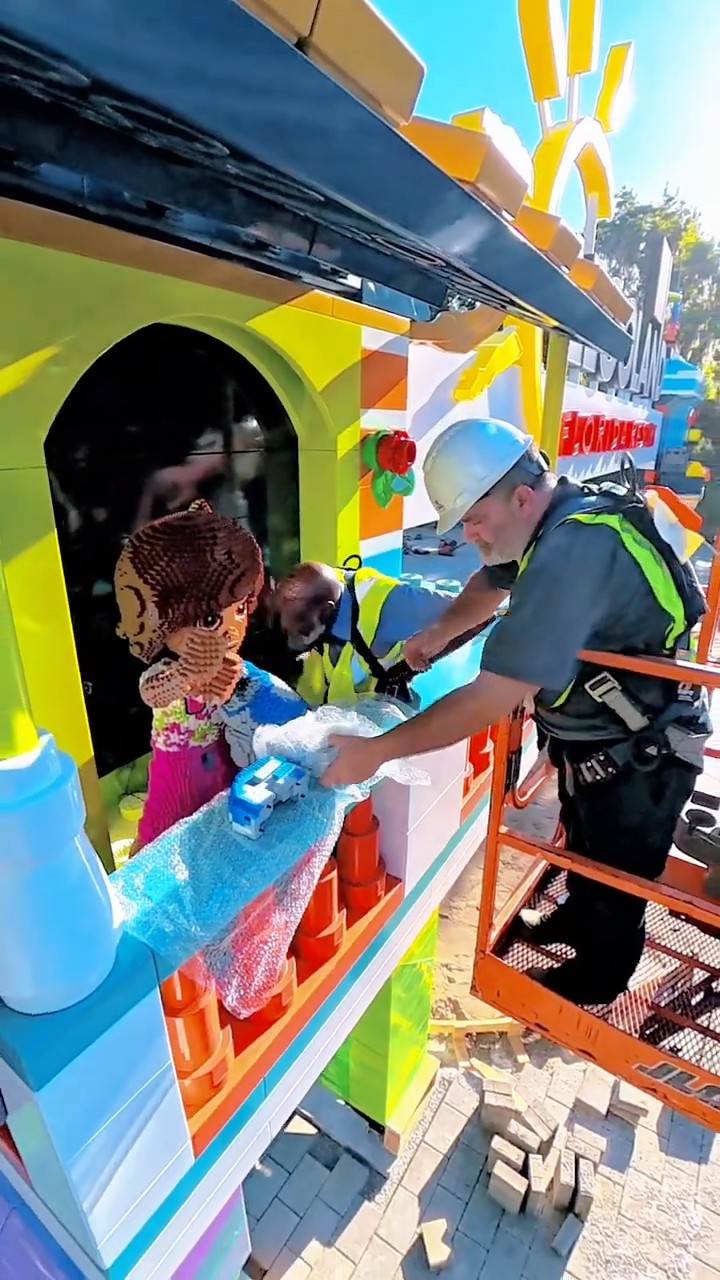 A worker in a white hard hat and a high visibility harness stands on an orange lift platform, peeling bubble wrap away from a large brick-built figure with brown hair, against the brightly colored panels and green arch of the new portal. A second worker in a yellow high visibility vest is at work just behind him.