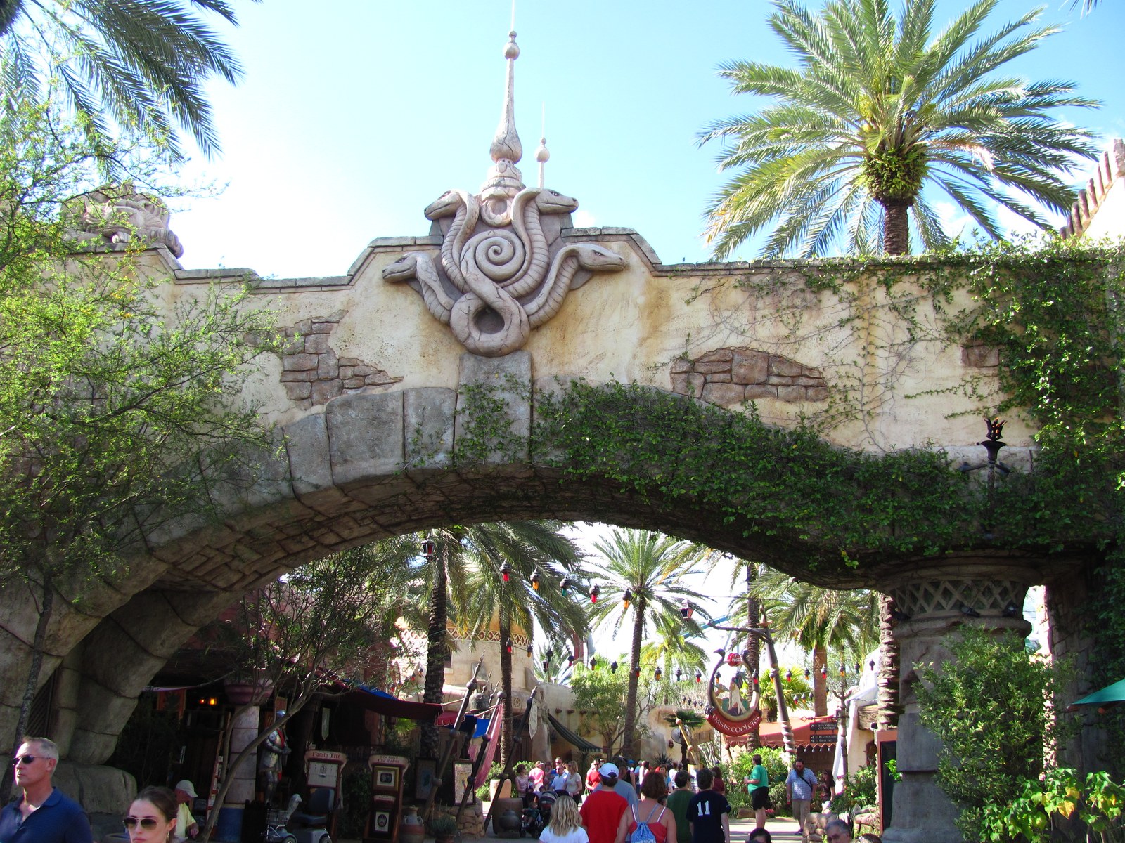 The ivy covered stone archway into The Lost Continent at Universal Islands of Adventure, carrying a carved medallion of snake heads coiled around a spiral at its crown with a tall finial spire above. Palm trees, hanging red, green and amber lanterns, market stalls and crowds of guests are visible through the arch, with a sign reading Oasis Coolers on the right.