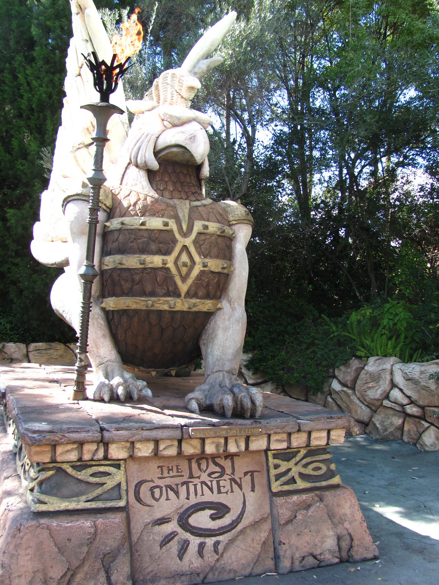 A white winged griffin statue with a brown scaled breast under gold studded armor straps sits on a stone plinth at the entrance to The Lost Continent at Universal Islands of Adventure. A lit flame torch on a tall metal stand stands beside it, the words THE LOST CONTINENT are carved into the plinth above a spiral emblem, and a small PLEASE DO NOT CLIMB plate runs along the base. Trees and rockwork surround the plaza.