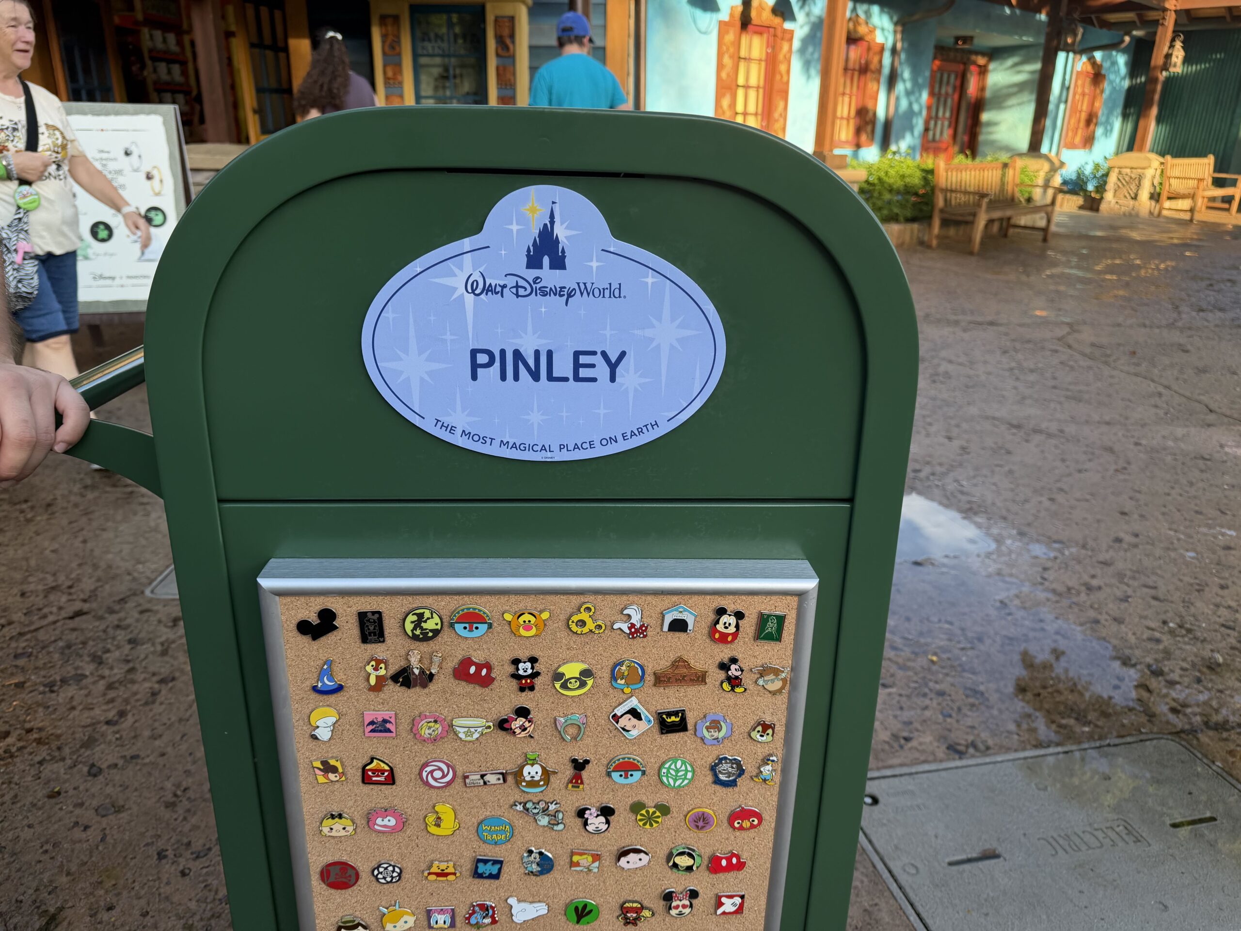 Close view of a green Disney trash can. An oversized oval Cast Member nametag on the upper front shows Cinderella Castle above the Walt Disney World logo, the name PINLEY in large letters, and the line The Most Magical Place on Earth. Below it, a cork board in a brushed metal frame is covered in rows of Disney trading pins.