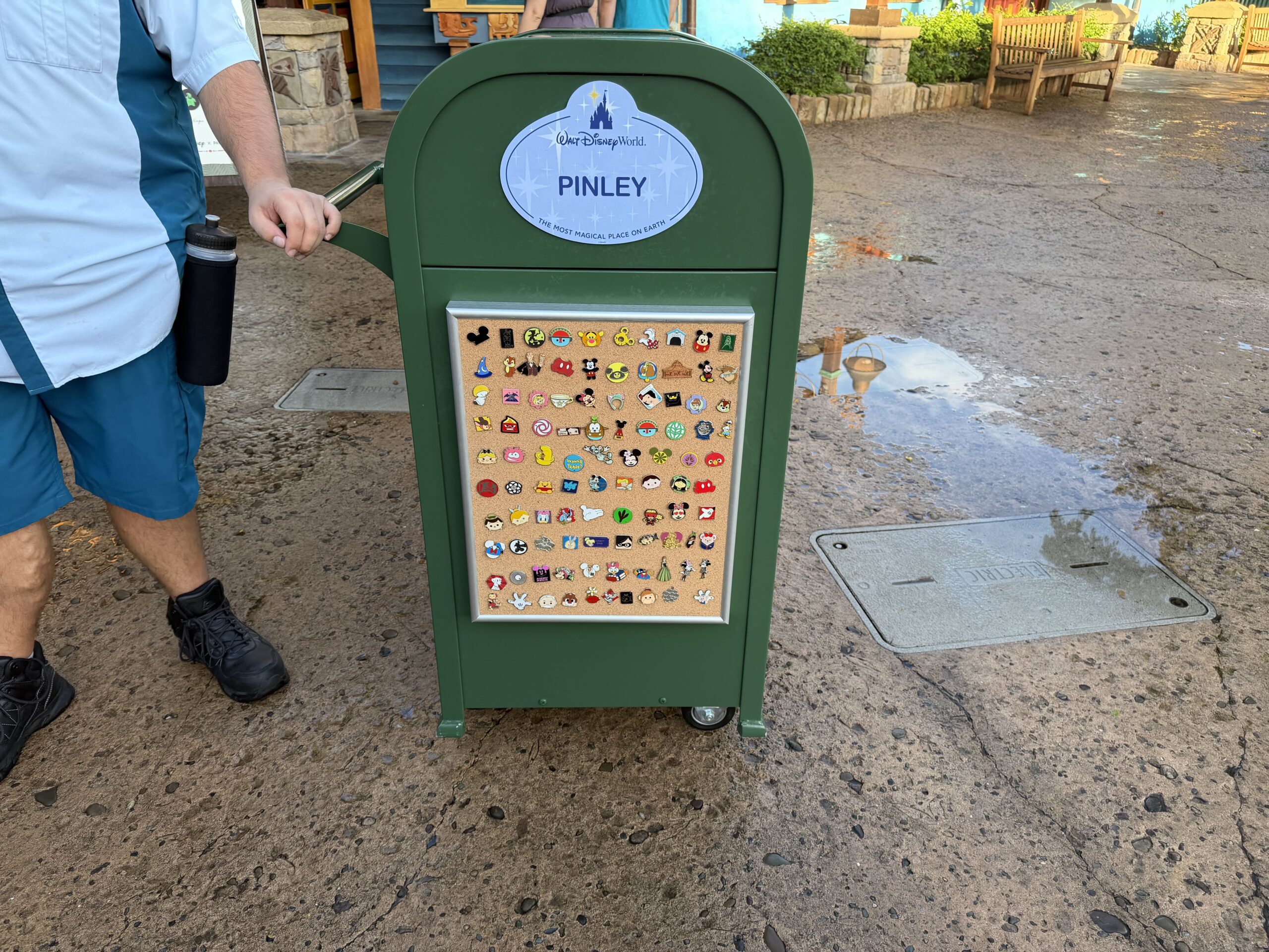 A Cast Member in a light blue shirt and teal shorts stands beside a green Disney trash can with a hand on its long metal push handle, on a damp concrete walkway on Discovery Island. A small wheel shows at the base of the can, the PINLEY nametag near the top and a cork board of trading pins below. Wooden benches and colorful Discovery Island buildings are in the background.